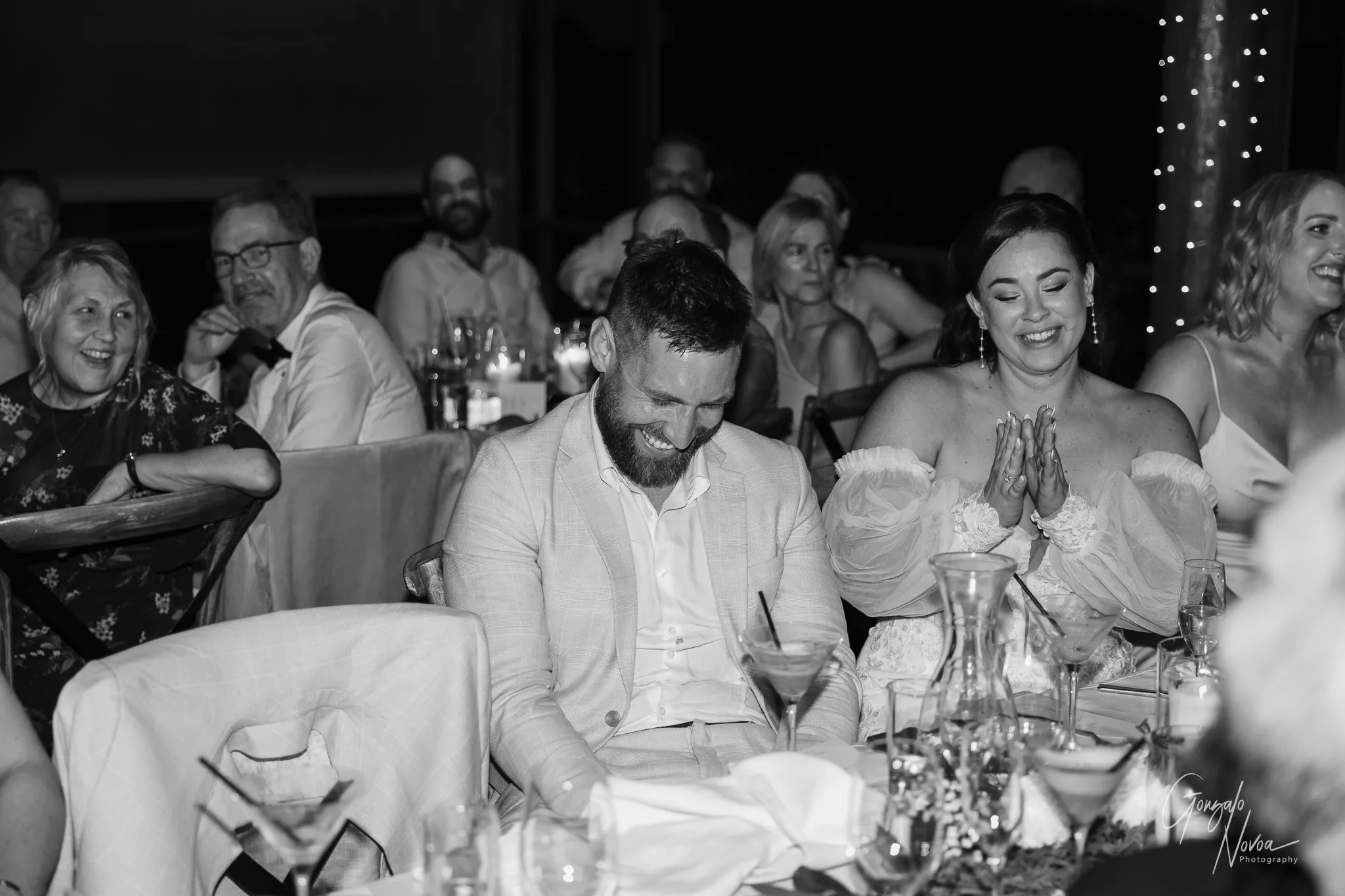 The groom bends forward laughing, while the bride smiles and claps her hands during a heartfelt speech at their wedding reception. Guests around them are laughing and smiling warmly. Fairy lights and candles create a cosy, festive atmosphere, and drinks and glassware are visible on the table.