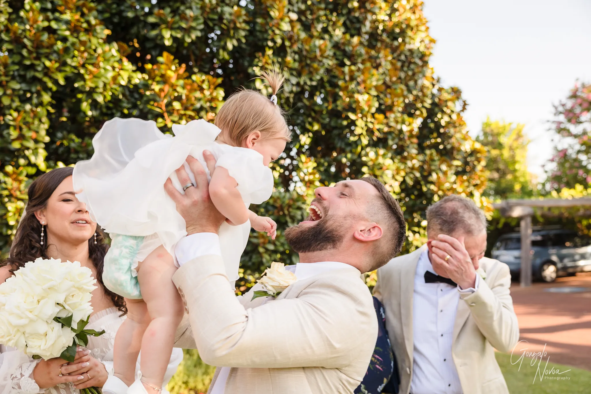 Groom James laughing joyfully while lifting his daughter Arlea in the air, with bride Tahnee smiling beside him and a guest in the background wiping his eyes