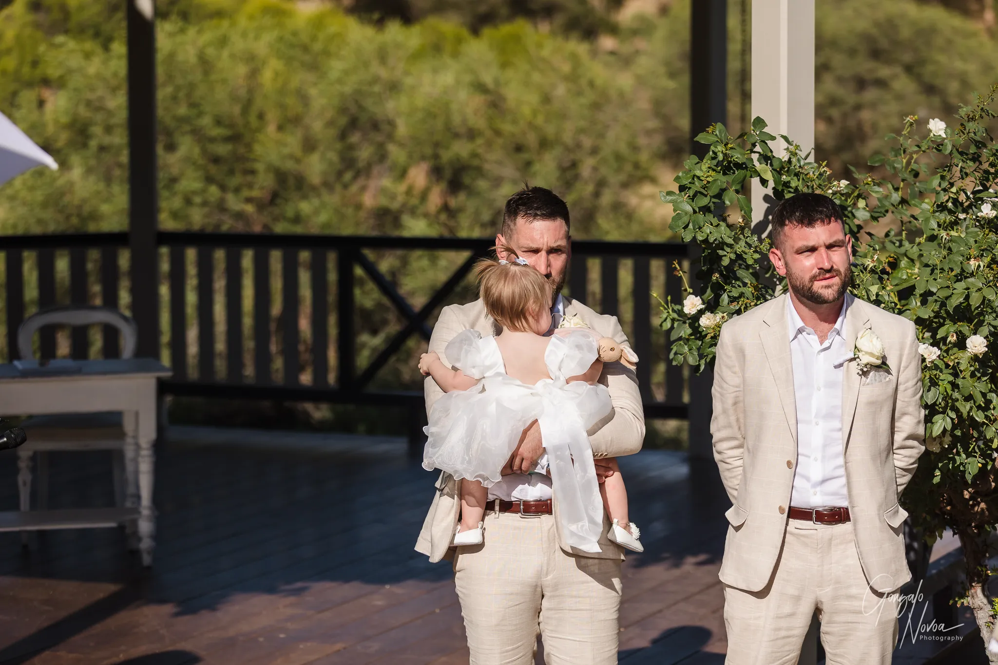 James holds his daughter Arlea in his arms at the altar, overcome with emotion as he sees Tahnee approaching. A groomsman stands beside them, smiling warmly against the backdrop of the garden ceremony at Sittella Winery.