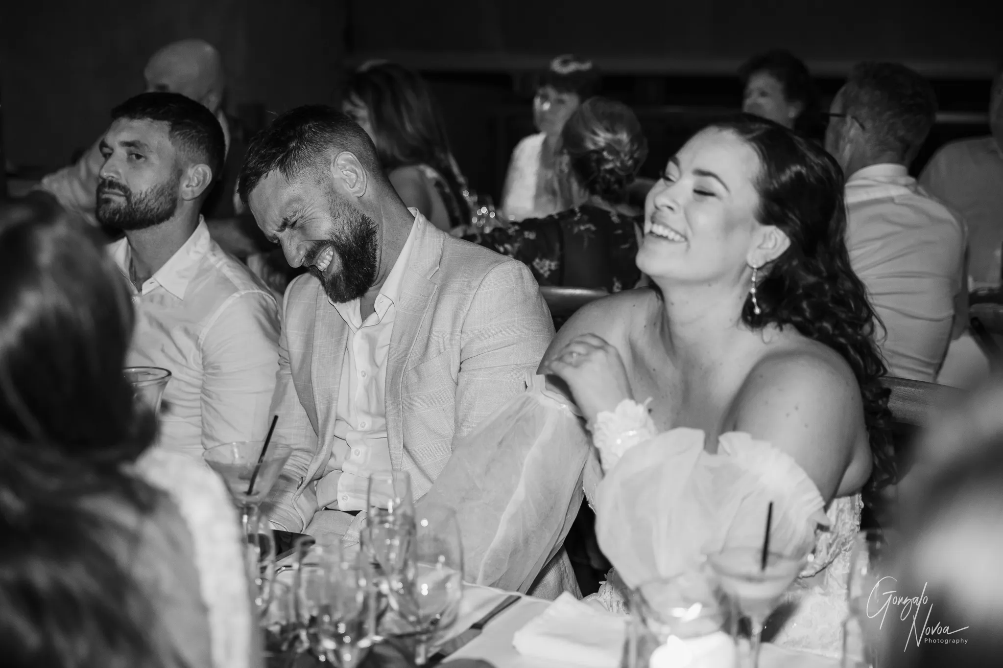 The groom and bride burst into laughter during a wedding reception speech. The groom leans forward, laughing with his eyes closed, while the bride smiles brightly beside him. Guests around them are also smiling and enjoying the moment, and glasses and candles fill the table in front of them.