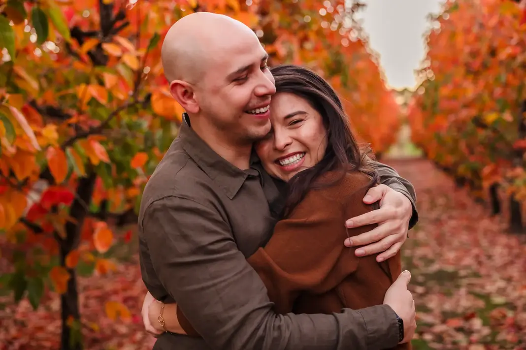 Luis and Sandra embrace joyfully among rows of autumn trees, both beaming with huge smiles after their surprise proposal.
