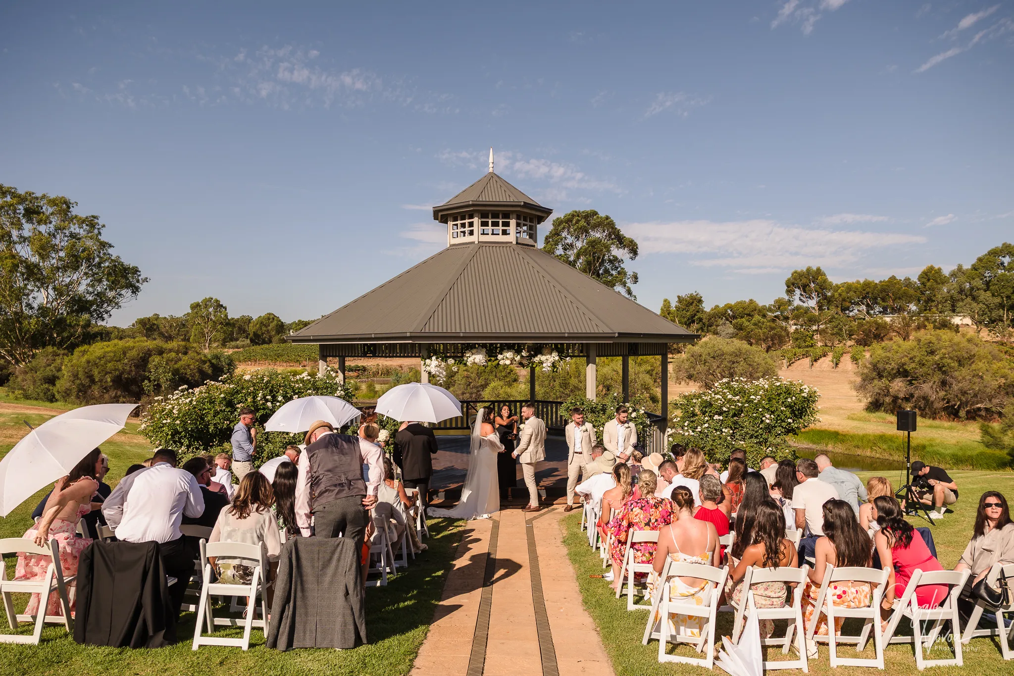 Wide view of Tahnee and James standing at the altar under the gazebo at Sittella Winery, holding hands during their ceremony while guests, some using white umbrellas for shade, watch from their seats on a sunny afternoon.