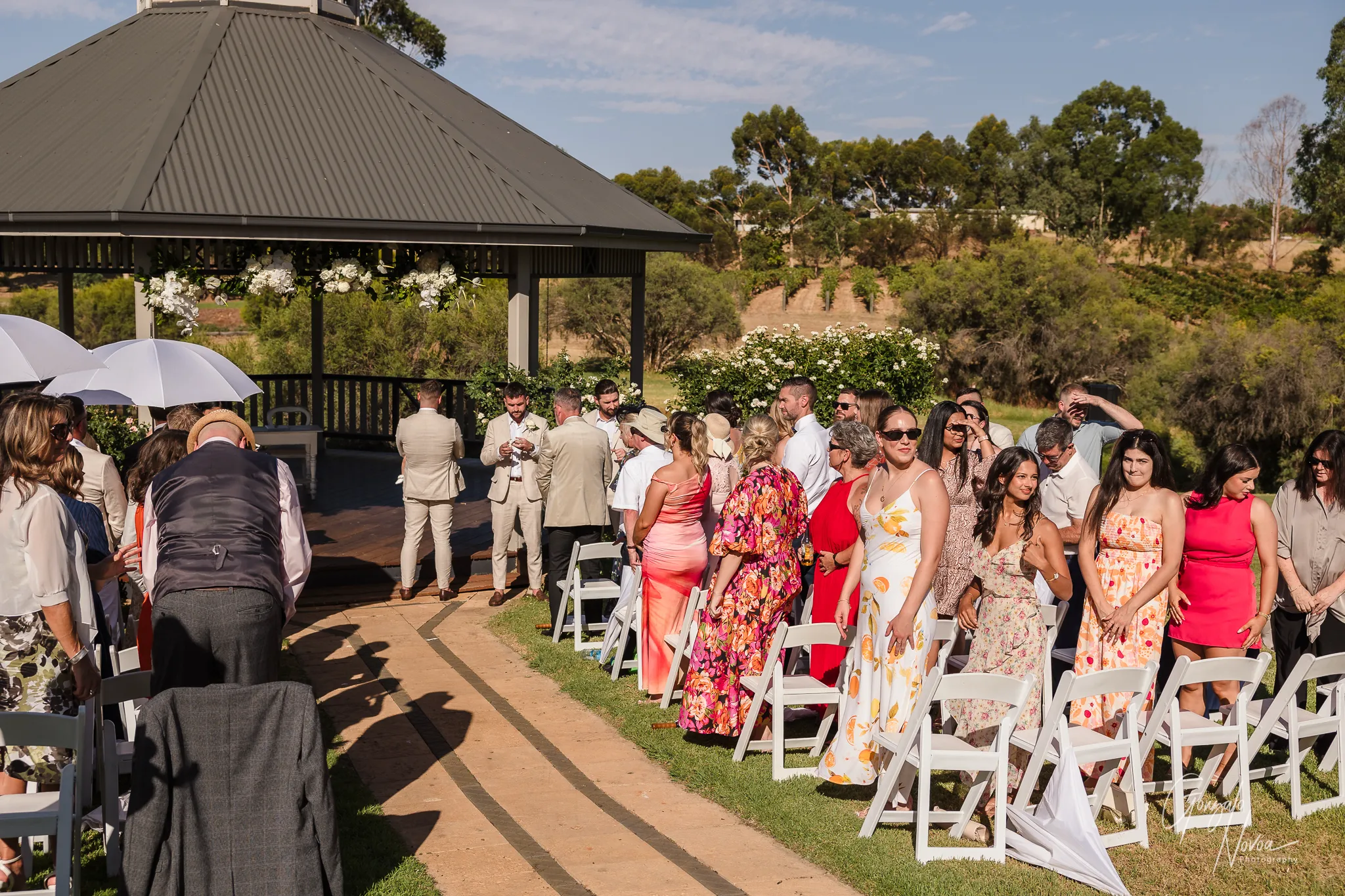 James turns his back to the aisle at Sittella Winery, waiting to see Tahnee until she walks closer. Guests stand and watch as the bride begins her long walk under the vineyard archway toward the flower-decorated gazebo.
