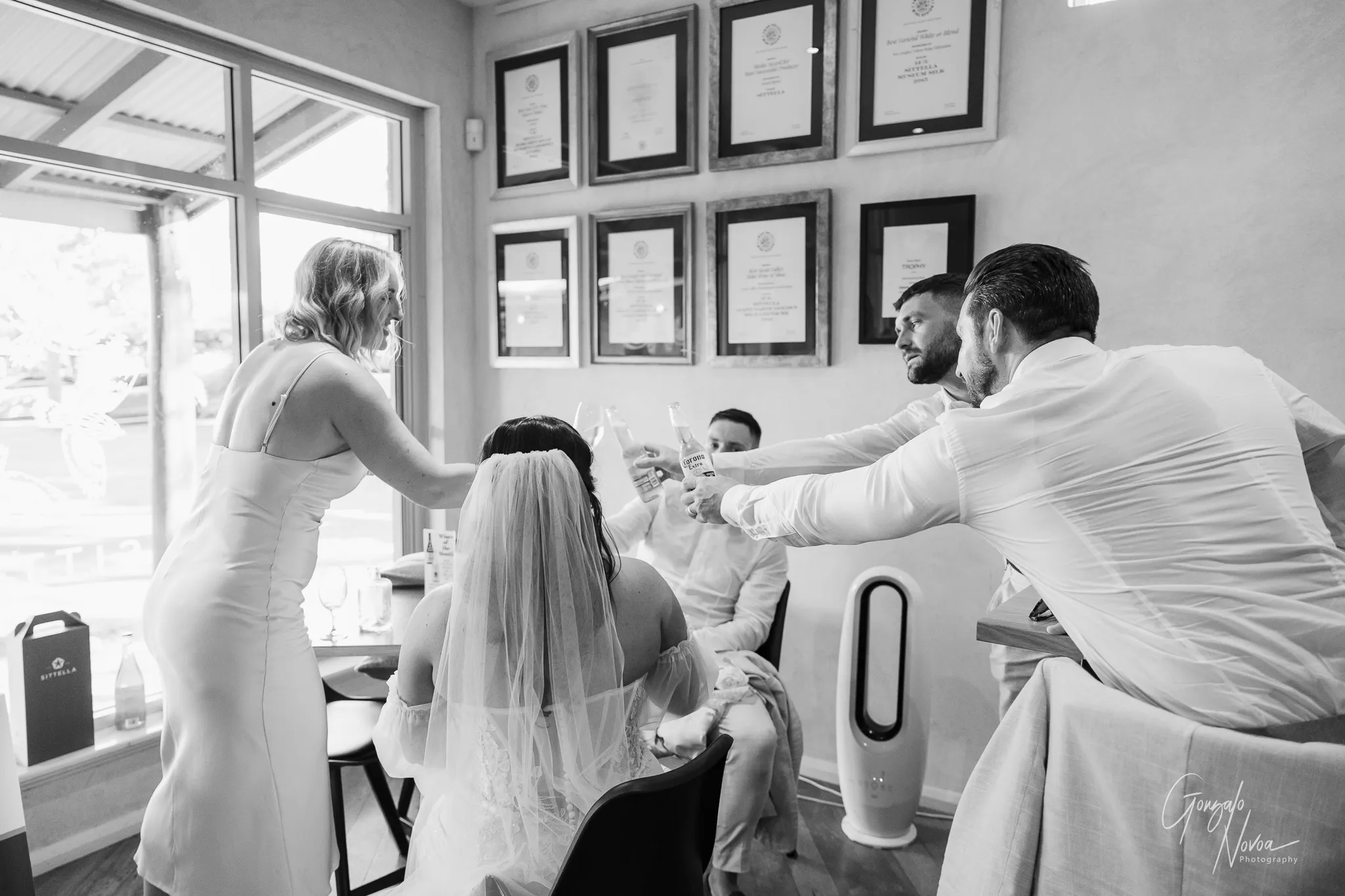 Black and white image of Tahnee and James toasting with their bridal party while seated inside the Sittella Winery cellar door, surrounded by framed wine awards on the wall