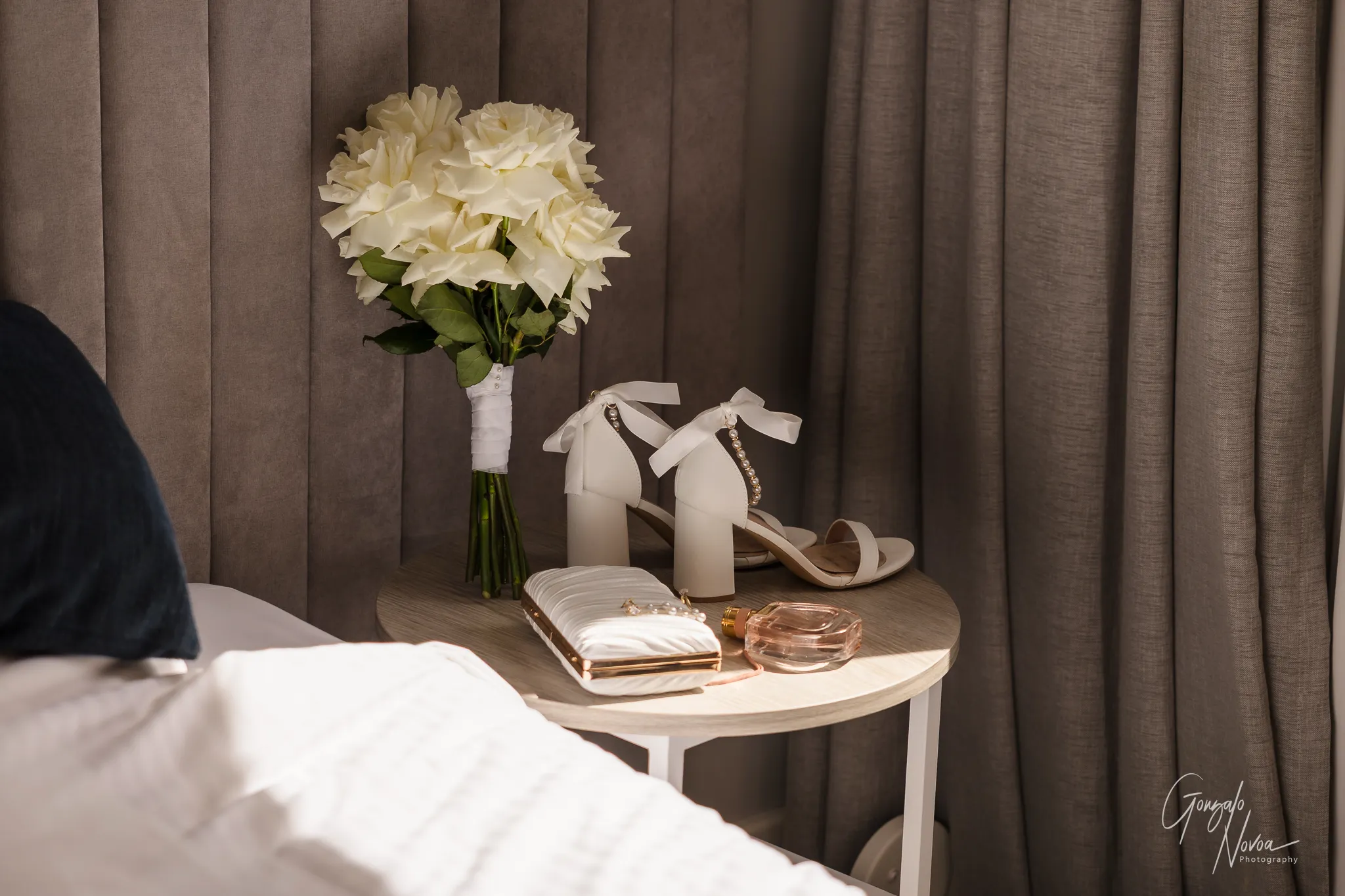 Bridal flat lay featuring Tahnee’s white rose bouquet, pearl-accented heels, a white clutch and a bottle of perfume placed neatly on a small round table by a bed. Natural light softly illuminates the scene, highlighting the elegant, romantic details before the wedding day.