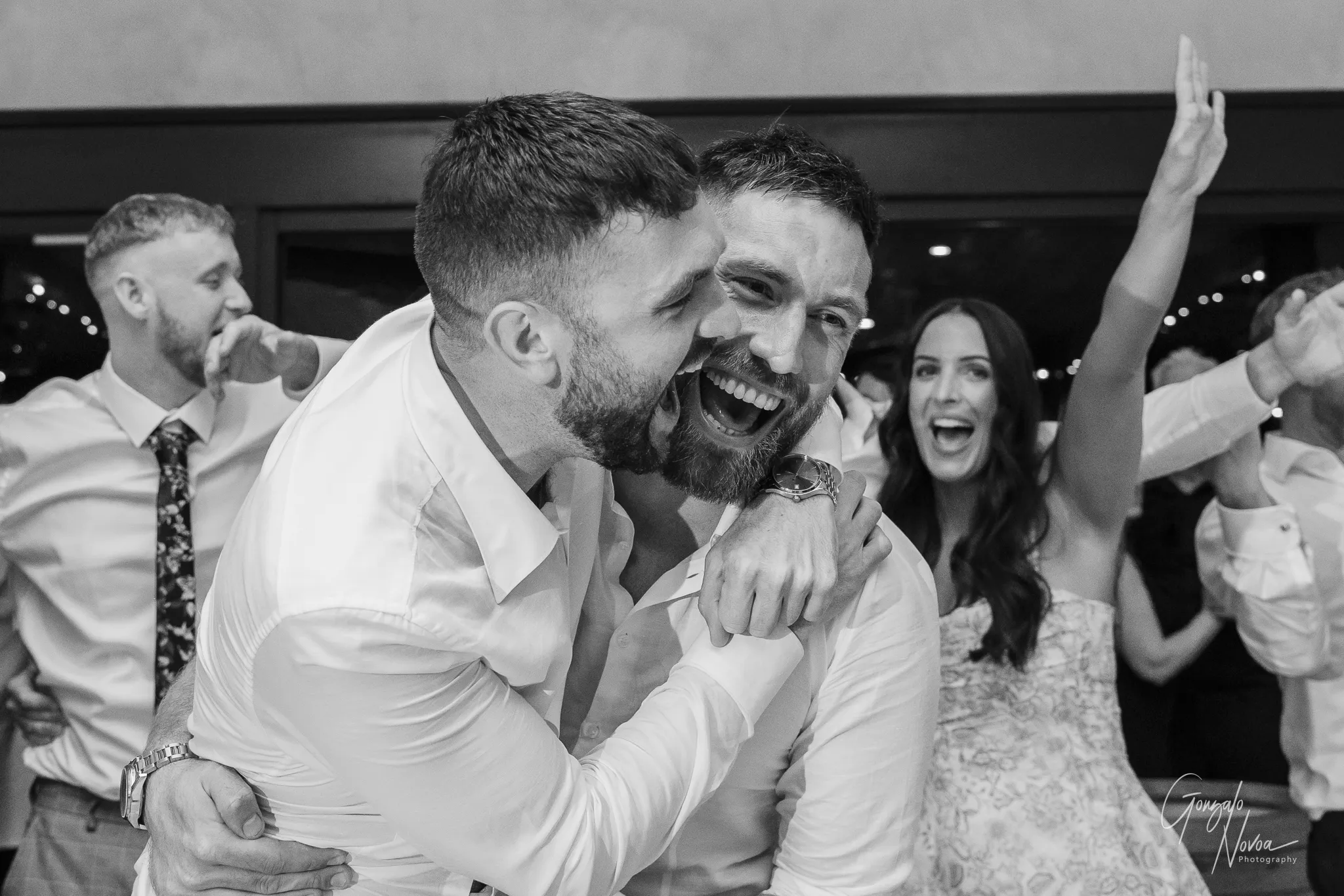 The groom and his brother laugh uncontrollably on the dance floor during the wedding reception. The groom, wearing a white open-collar shirt, smiles widely as his brother playfully pretends to bite his cheek in a fun, energetic hug. Both are clearly having the time of their lives. In the background, a woman in a patterned strapless dress cheers with her arms raised, adding to the lively atmosphere.