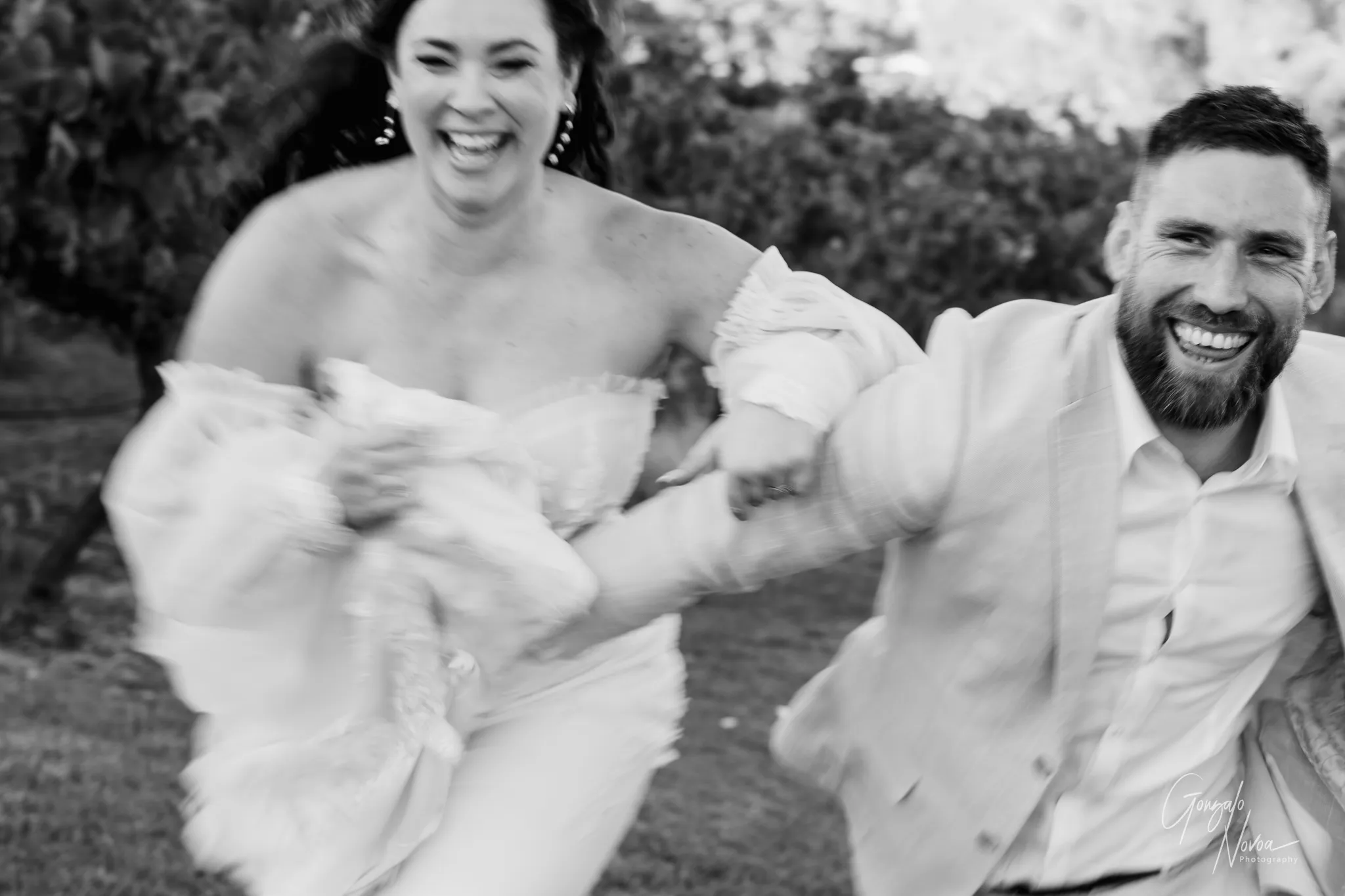 Blurry black and white photo of a laughing bride and groom running hand in hand across a vineyard, both smiling brightly and full of joy