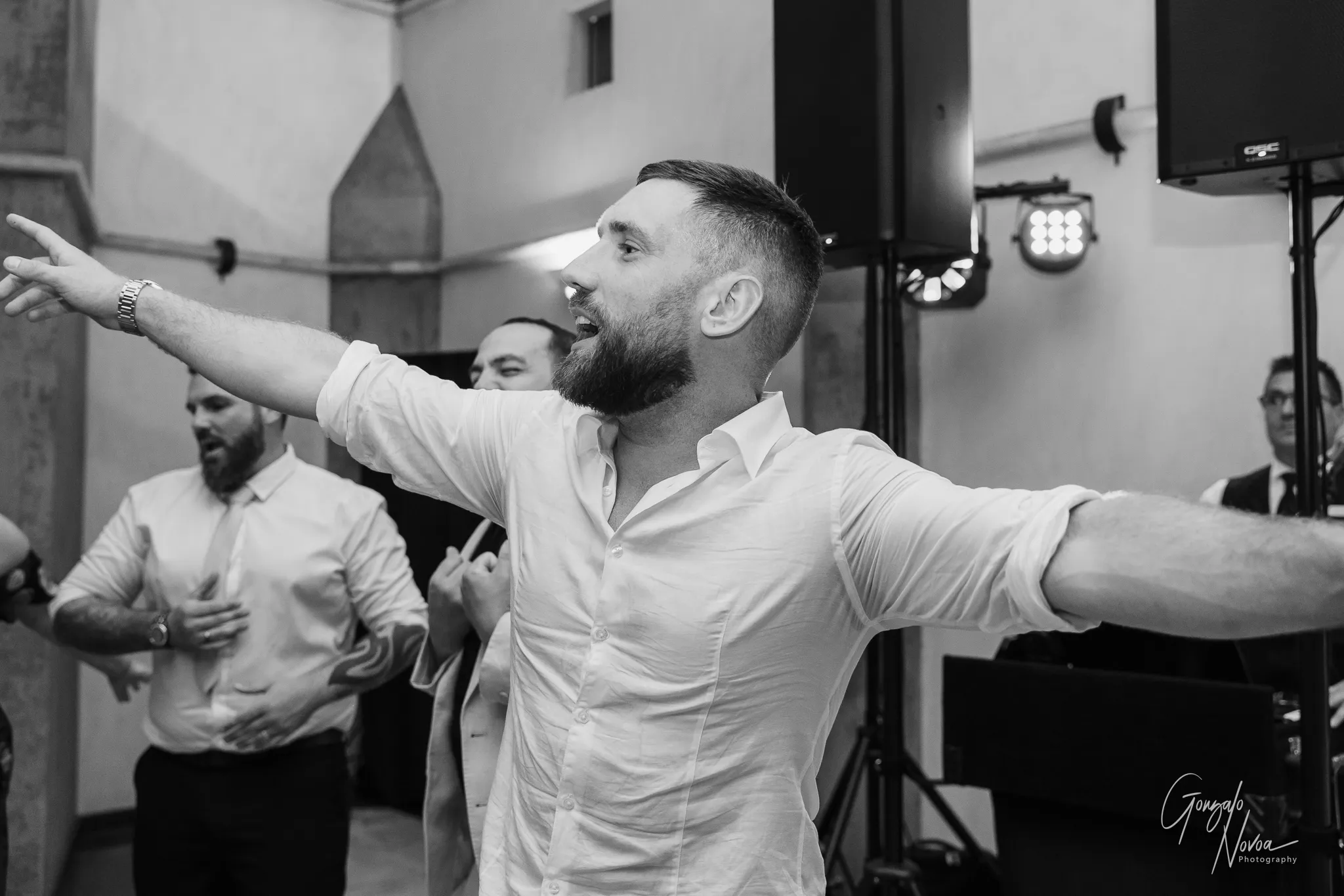 The groom, in a white shirt with rolled-up sleeves, points outward and sings passionately with a big smile on his face during the dance floor celebrations. Guests cheer around him.