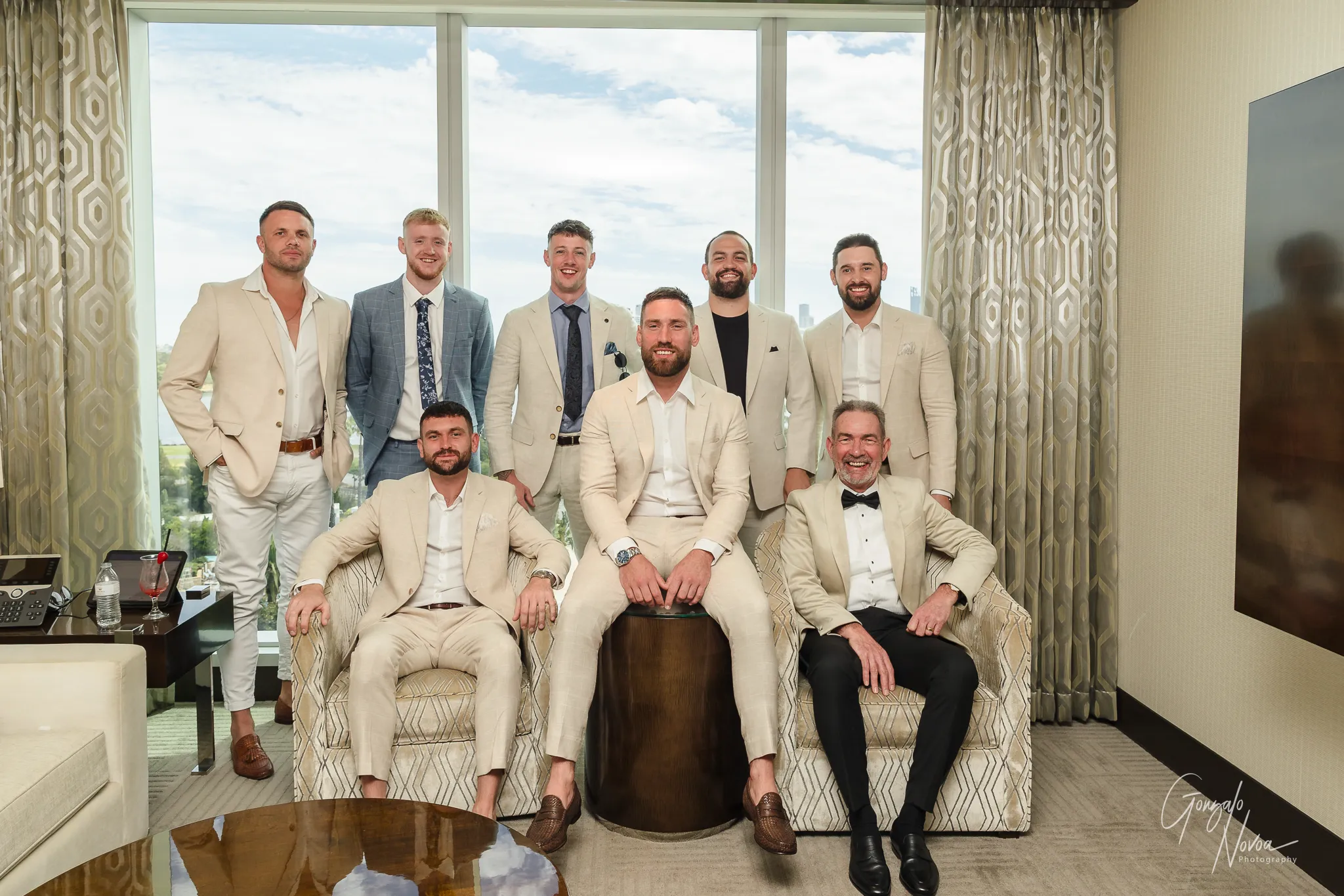 Colour photo of James seated with his groomsmen, friends and his father in a bright Crown Towers Perth hotel room. Everyone is dressed in coordinating beige and cream suits, smiling confidently with a city skyline visible through the large windows behind them.
