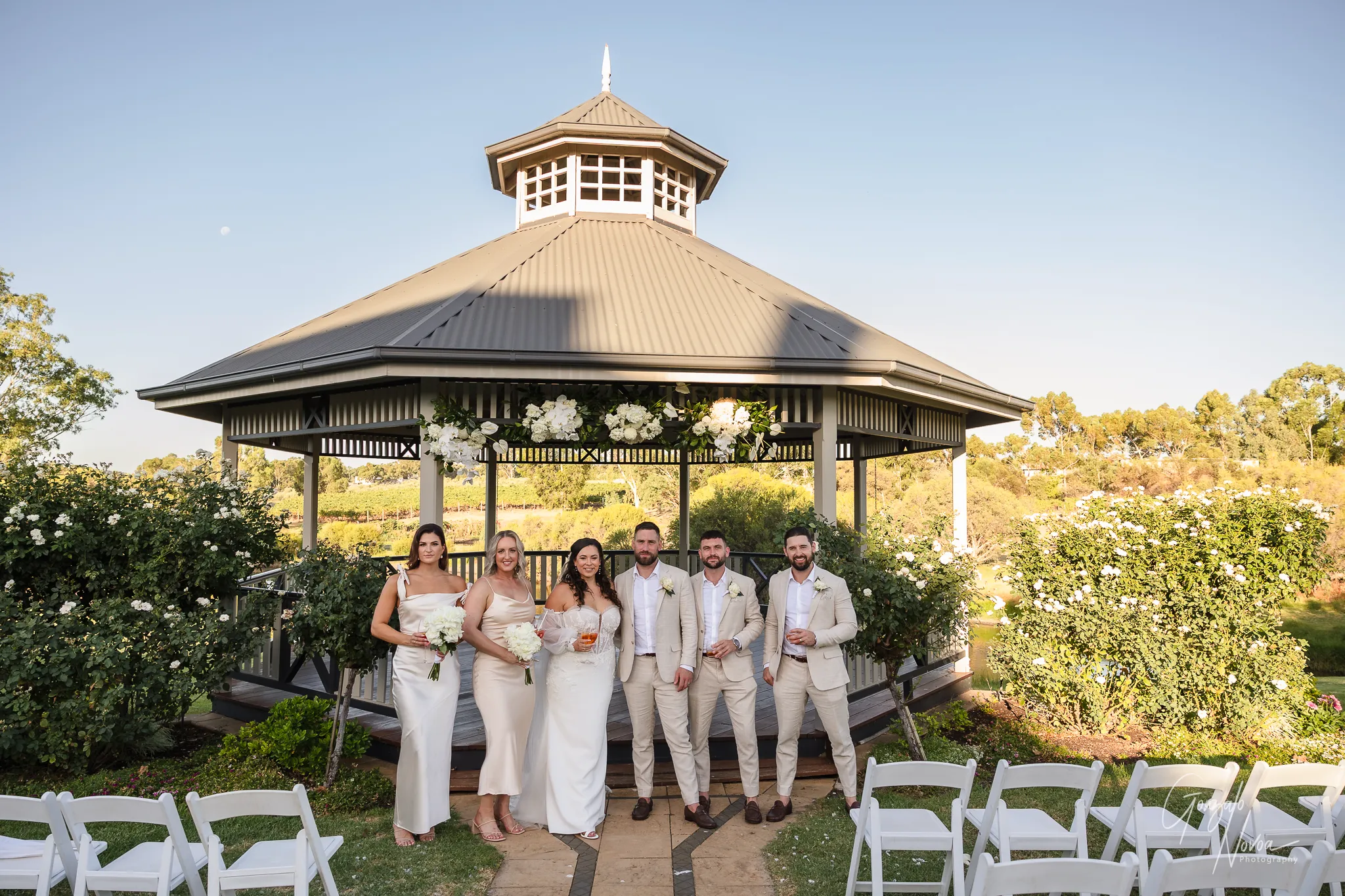 Tahnee, James, and their bridal party standing in front of the Sittella gazebo surrounded by white flowers and vineyards, smiling and relaxed after the ceremony