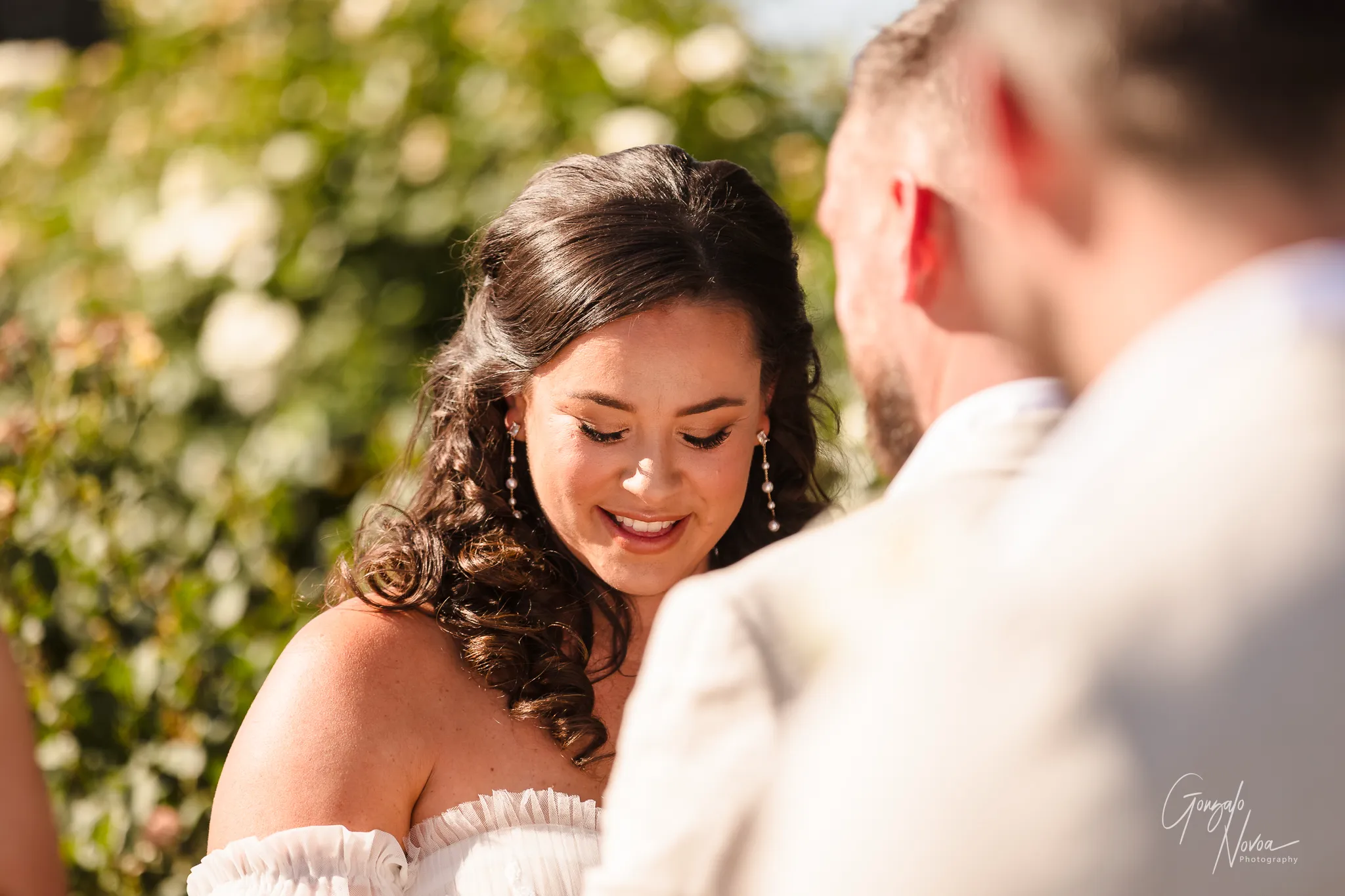 Close-up of bride Tahnee smiling tenderly while reading her vows during the wedding ceremony, with groom James slightly blurred in the foreground.