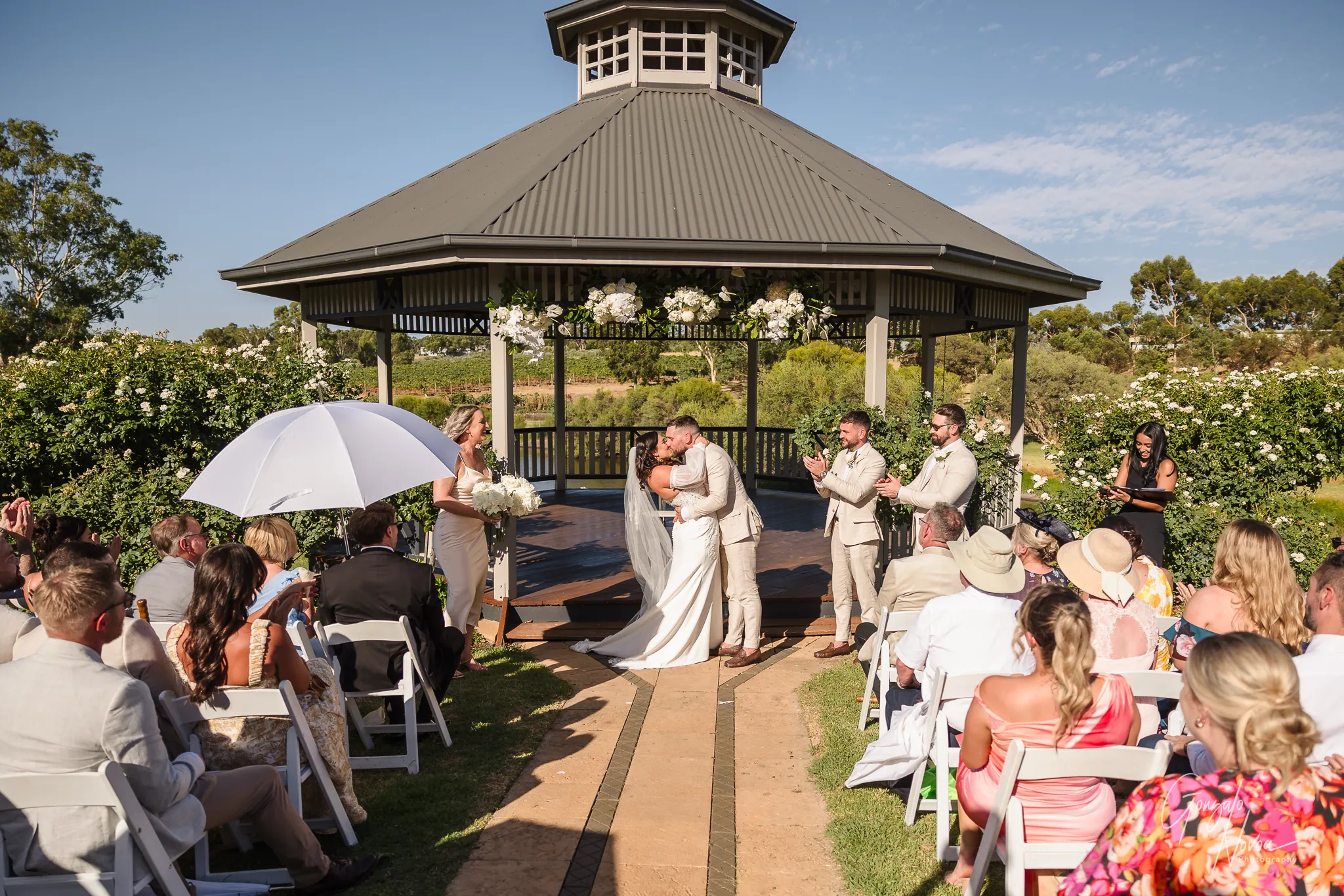 Bride and groom sharing their first kiss as a married couple under the flower-adorned gazebo at Sittella Winery, with their wedding party and guests applauding joyfully around them.