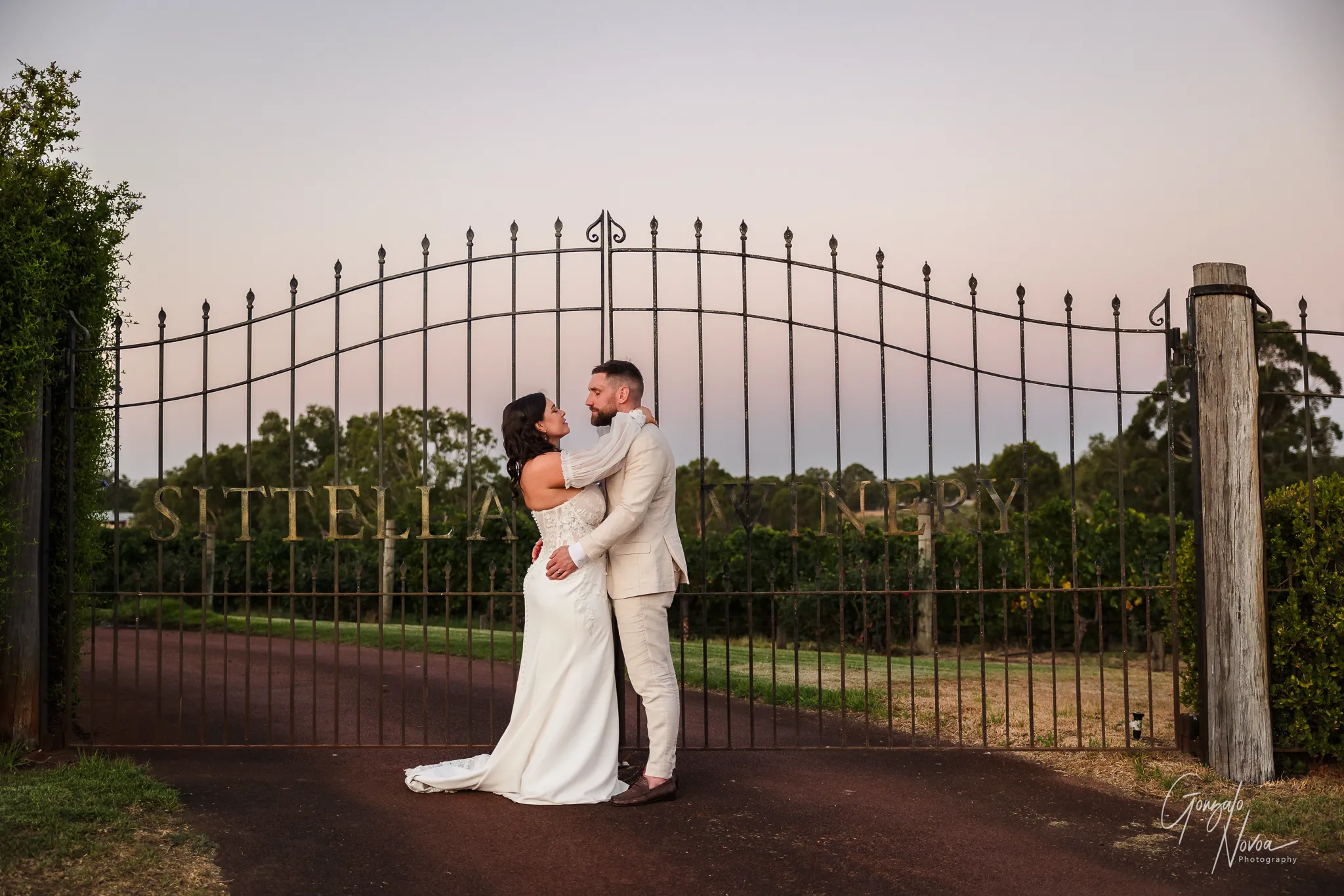 Bride and groom sharing an intimate embrace in front of the iron gates at Sittella Winery. The scene is captured in black and white, with the couple centred against the intricate gate design and lush vineyard landscape behind them.