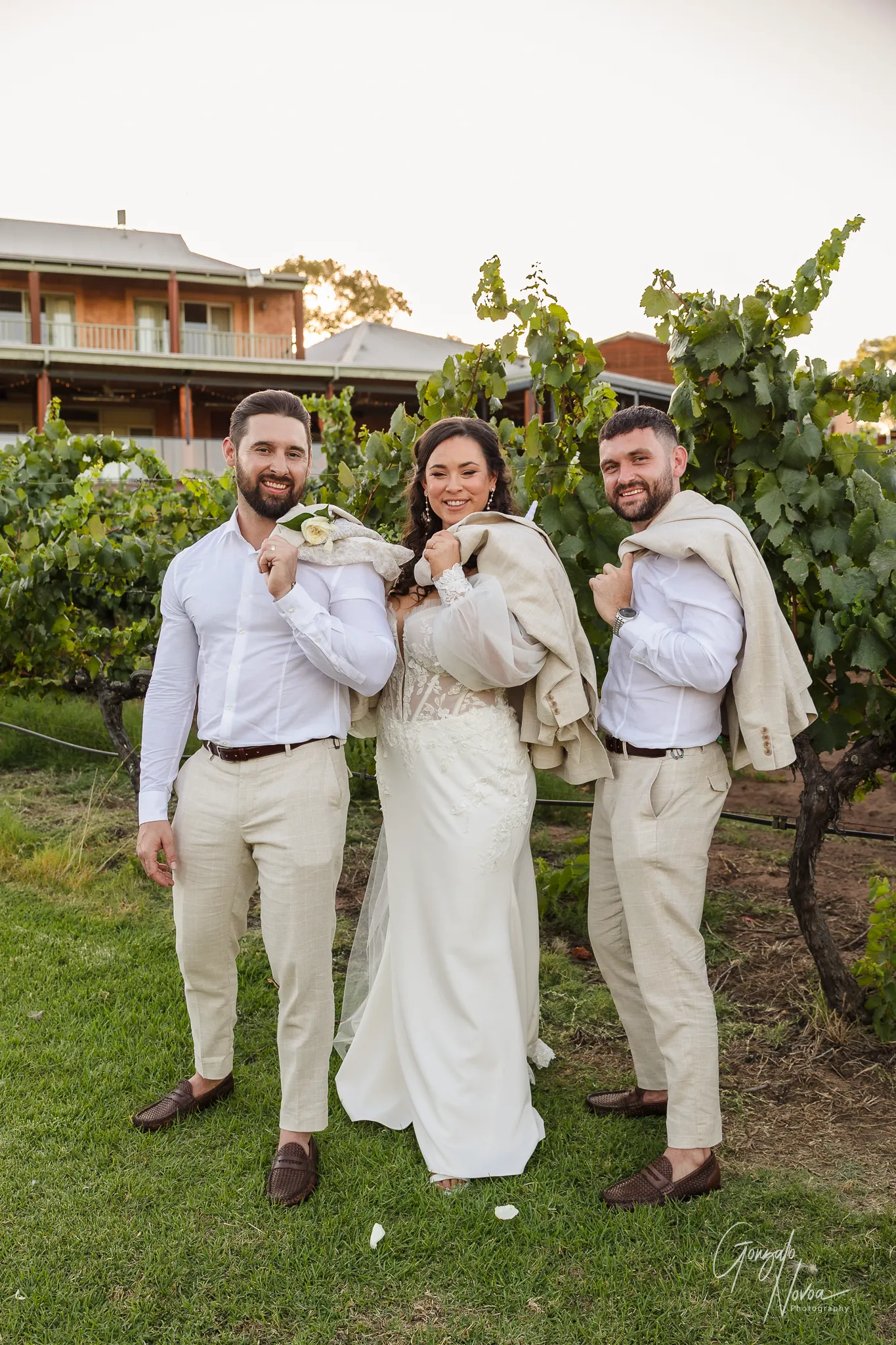 Bride Tahnee standing between two groomsmen in a vineyard, all three holding their jackets over their shoulders and smiling at the camera