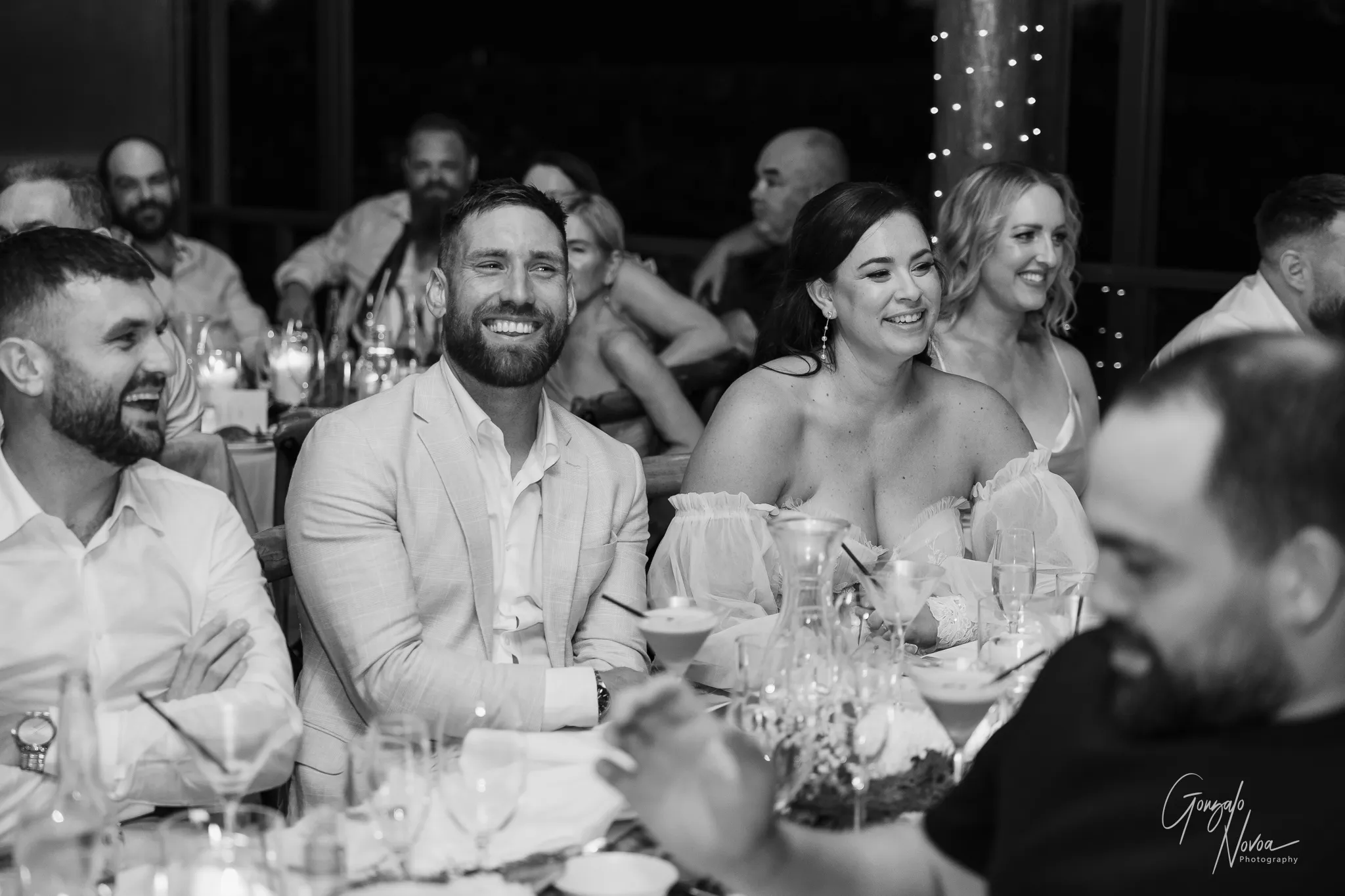The groom, bride, and guests laugh and smile during a wedding reception speech. The atmosphere is warm and lively, with fairy lights wrapped around a nearby column and candles glowing softly on the tables.