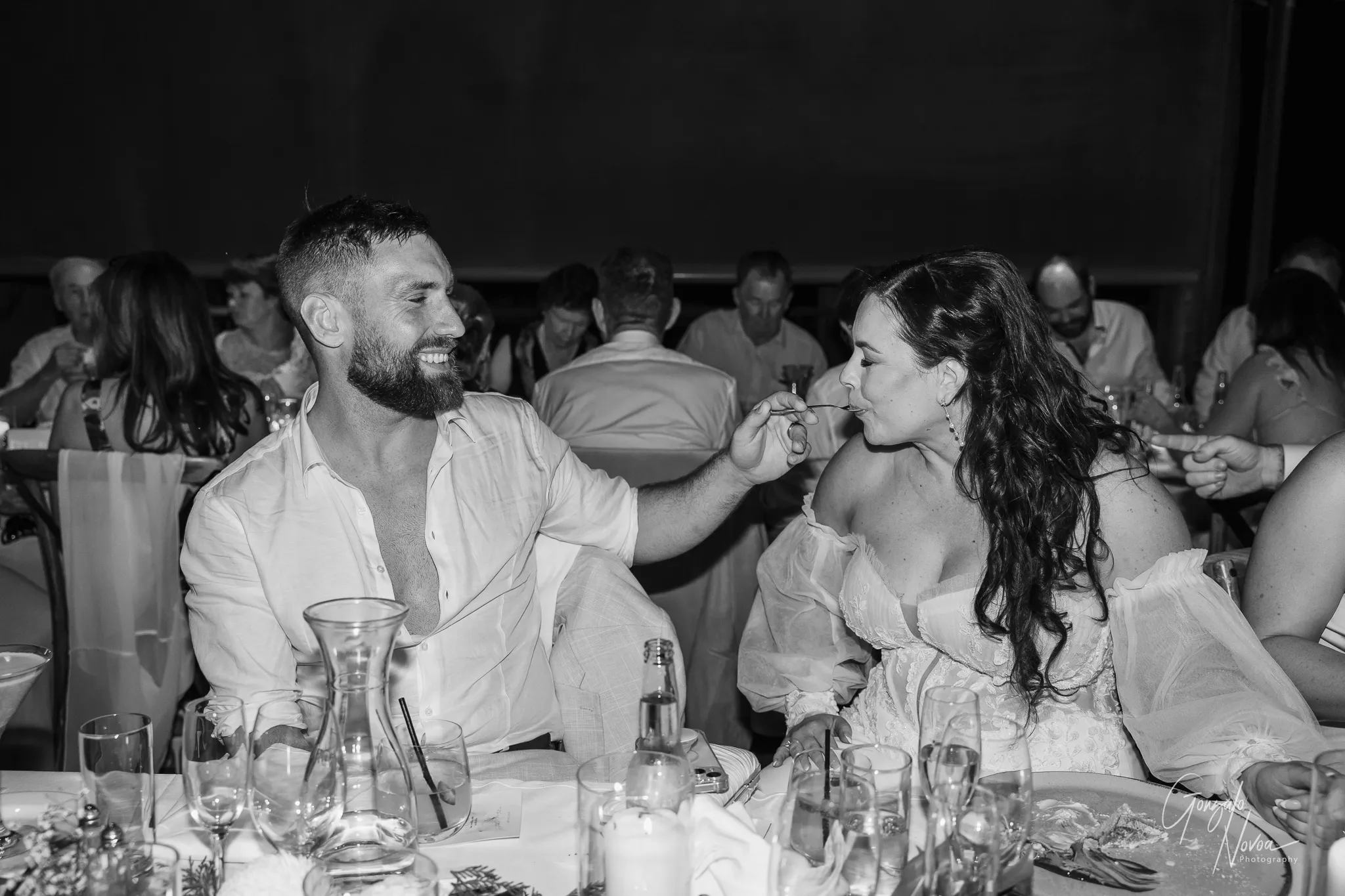 The groom, dressed in an open-collared white shirt, smiles warmly as he feeds the bride with a spoon during the reception. The bride, still in her beautiful off-shoulder lace gown, leans forward with a playful smile. They are surrounded by glasses, candles, and joyful guests at their wedding celebration, captured in a candid, affectionate moment.