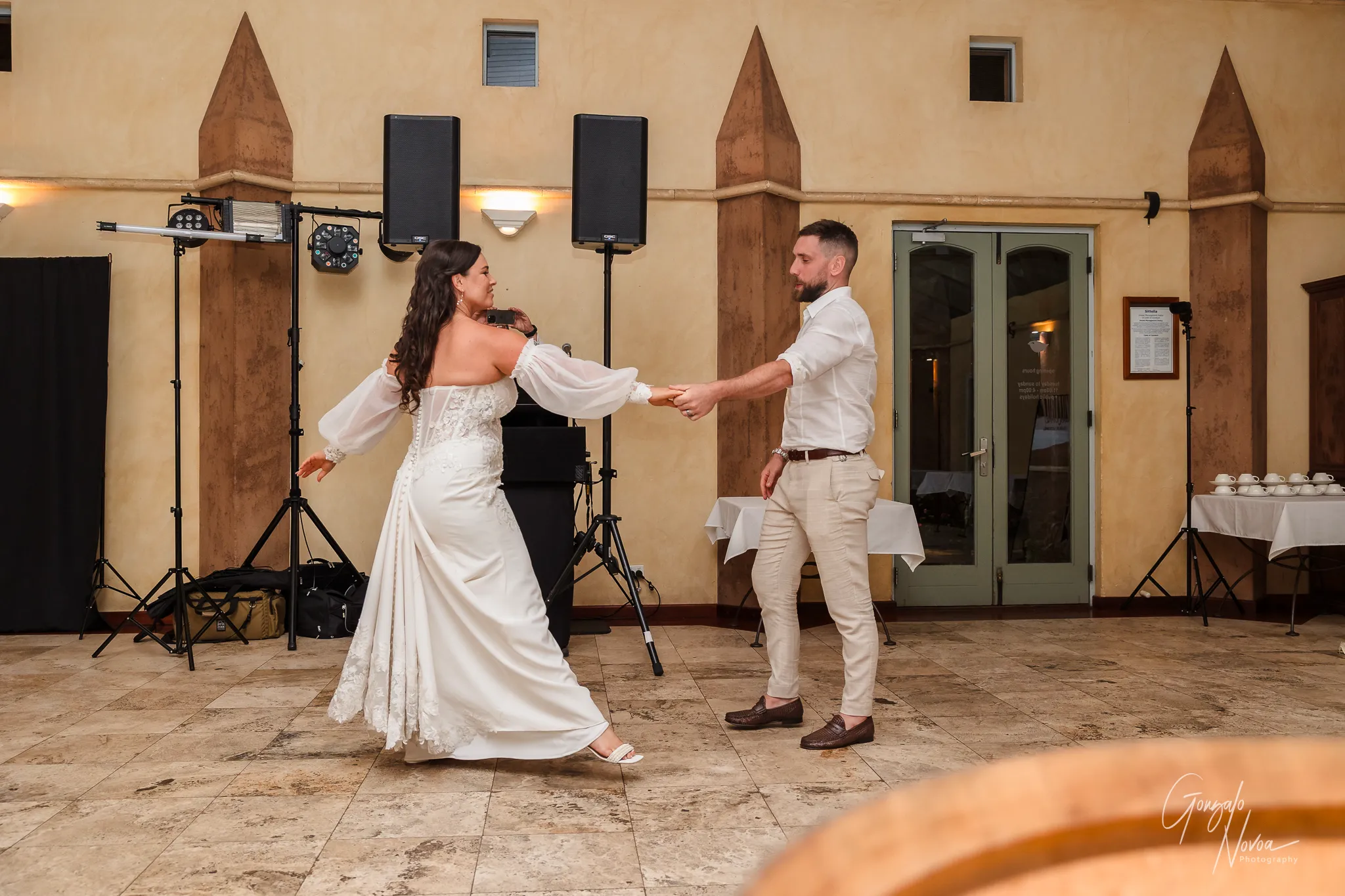 The bride and groom share their first dance at the reception, holding hands mid-twirl. The bride wears an off-shoulder gown with flowing sleeves and intricate lace details, while the groom is dressed casually in a white shirt and beige trousers. The background shows a simple indoor venue setup with speakers, lighting equipment, and tables adorned for the celebration.