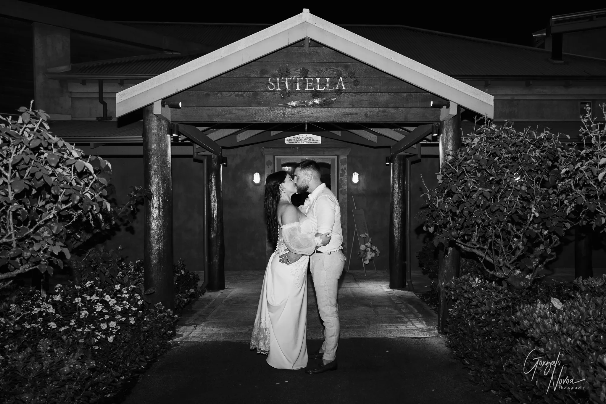 The bride and groom share a romantic kiss under the entrance archway at Sittella Winery. The black-and-white photo captures them holding each other close, surrounded by garden hedges and lit by soft exterior lights.