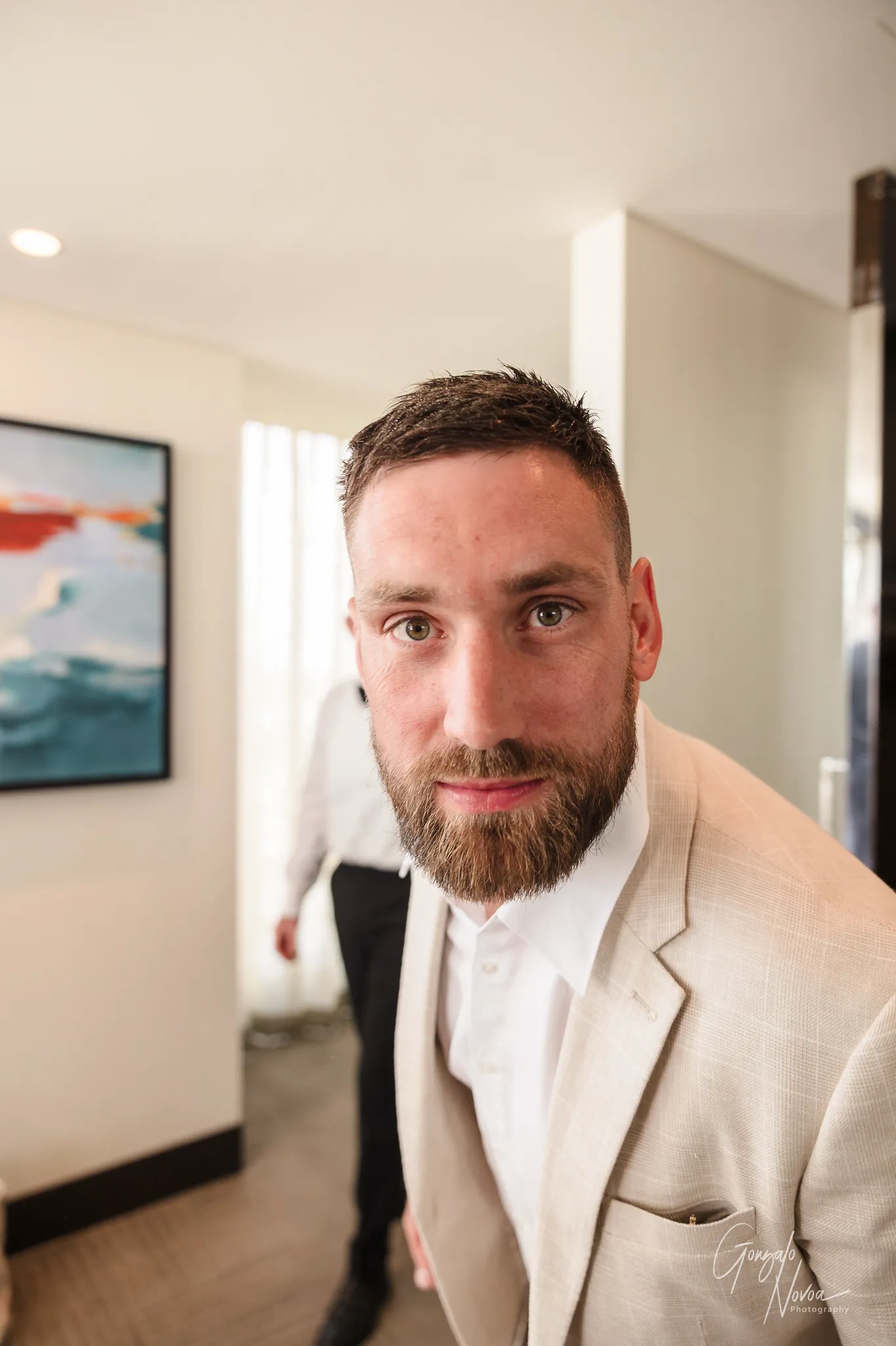 Close-up photo of James in a cream-coloured suit leaning playfully toward the camera inside a hotel room. He looks directly at the lens with a half-smile, full beard, and relaxed energy during the getting ready portion of the wedding day.
