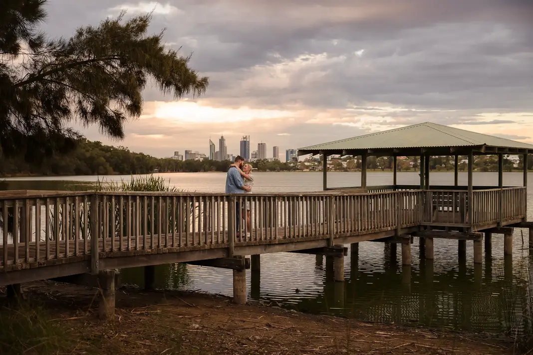 Wide-angle photo of Faith and Alex embracing on the boardwalk at Lake Monger, with trees, gazebo, water, and the Perth skyline in the background at sunset.