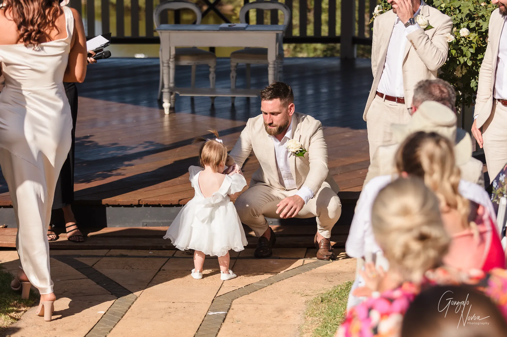 Groom James, dressed in a light beige suit, crouches down with a big smile to greet his daughter Arlea, who is making her way down the aisle in a white dress during the outdoor ceremony at Sittella Winery.