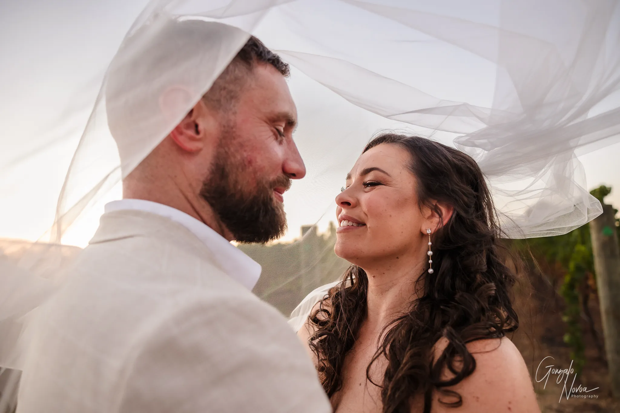 Close-up of the bride and groom smiling at each other, standing under the bride’s flowing veil during golden hour. The groom wears a light suit and the bride wears pearl earrings and a strapless lace wedding dress, surrounded by vineyard scenery.