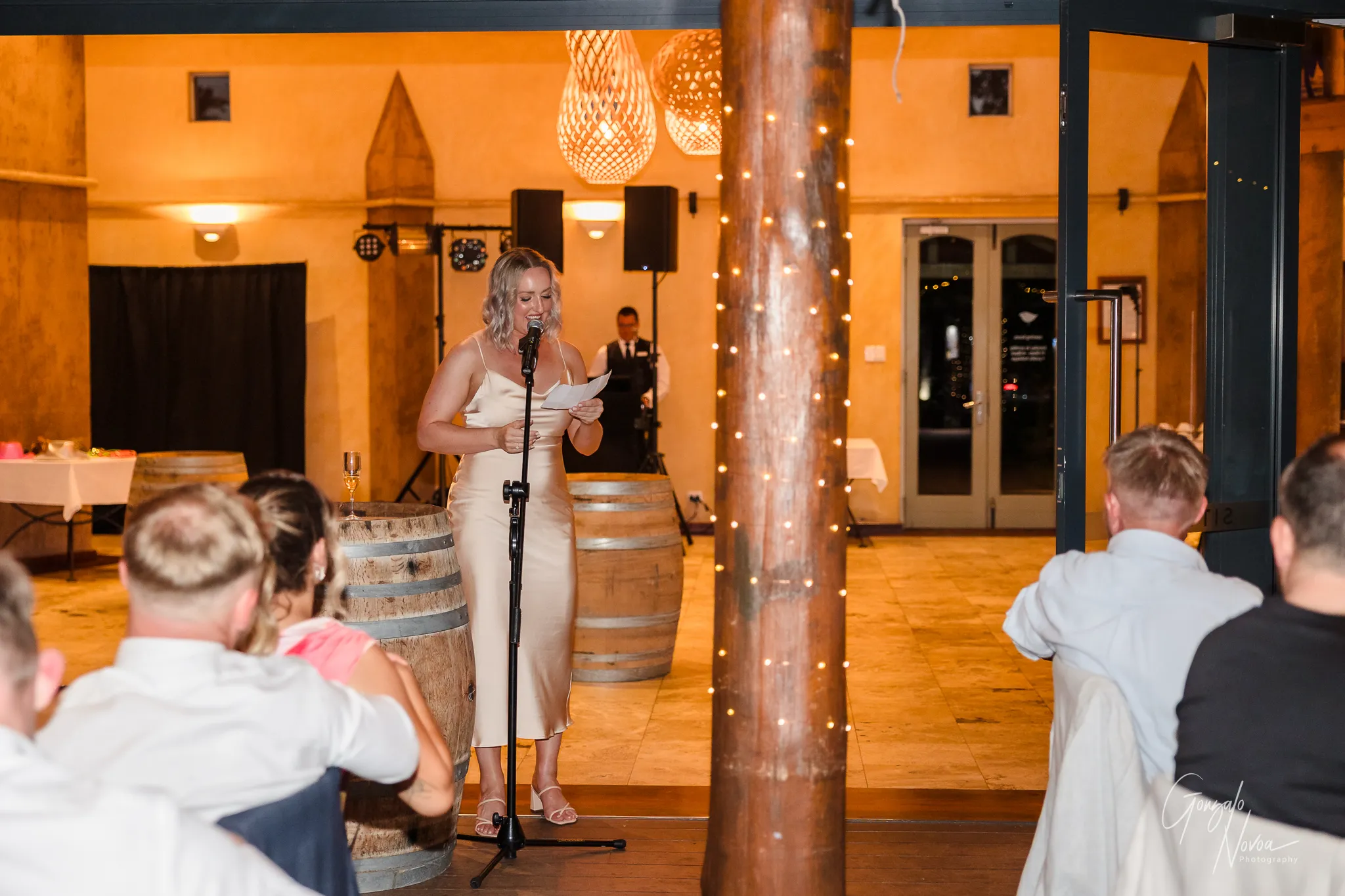 The Maid of Honour stands in front of a microphone, reading from her notes during her wedding speech. She wears a light satin gown and smiles as she speaks. The room is softly lit, with fairy lights wrapped around a wooden post and wine barrels visible in the background. Guests are seated, listening attentively.