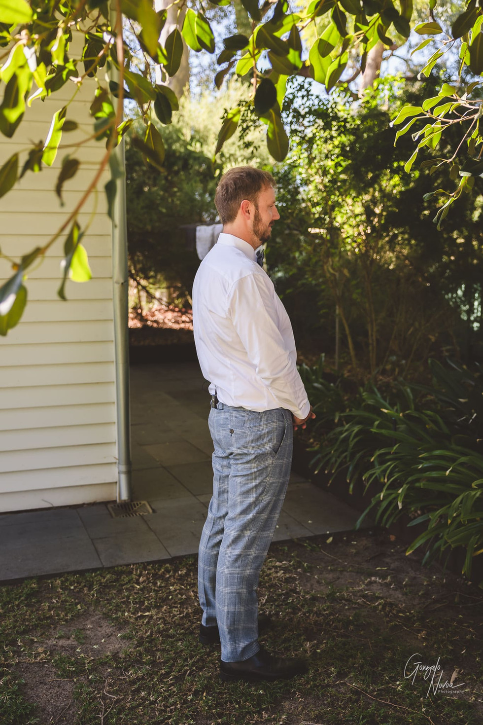 Groom stands in anticipation, waiting for his First Look with the bride, outdoors in a lush, garden setting.
