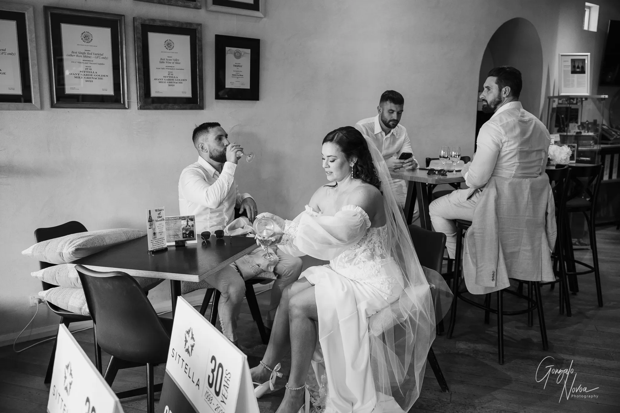 Black and white photo of Tahnee and James seated inside Sittella Winery’s cellar door, with Tahnee pouring a drink and James sipping from a wine glass, joined by groomsmen in the background