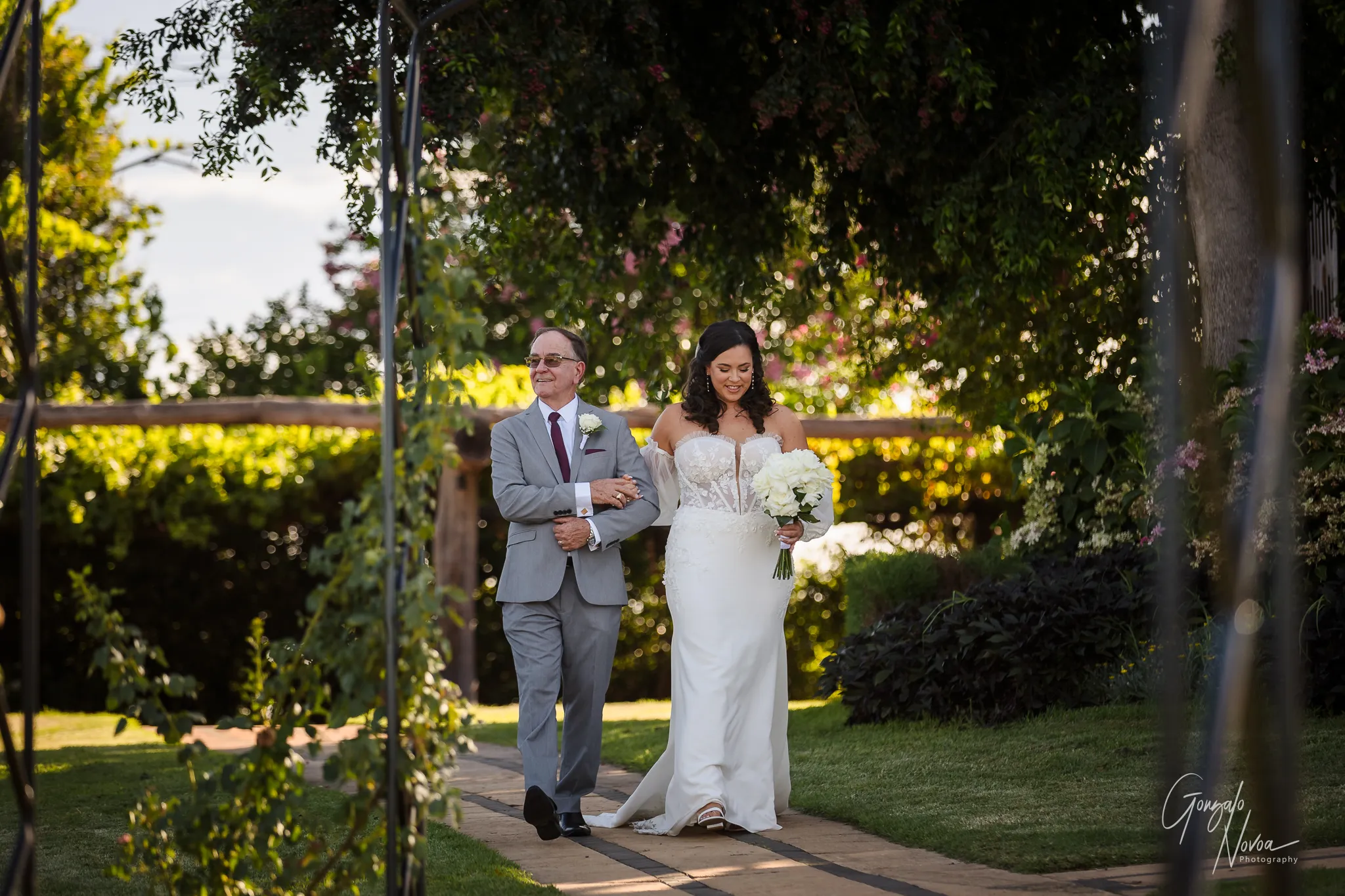 Tahnee walks arm-in-arm with her dad down the long garden path at Sittella Winery, surrounded by greenery and soft light. She carries a bouquet of white flowers and smiles as they approach the ceremony gazebo.