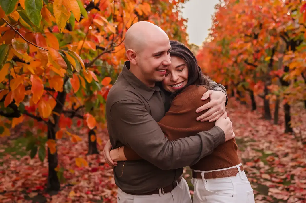 Luis and Sandra embrace with eyes closed and smiles on their faces, wrapped in each other’s arms among autumn trees after their engagement.