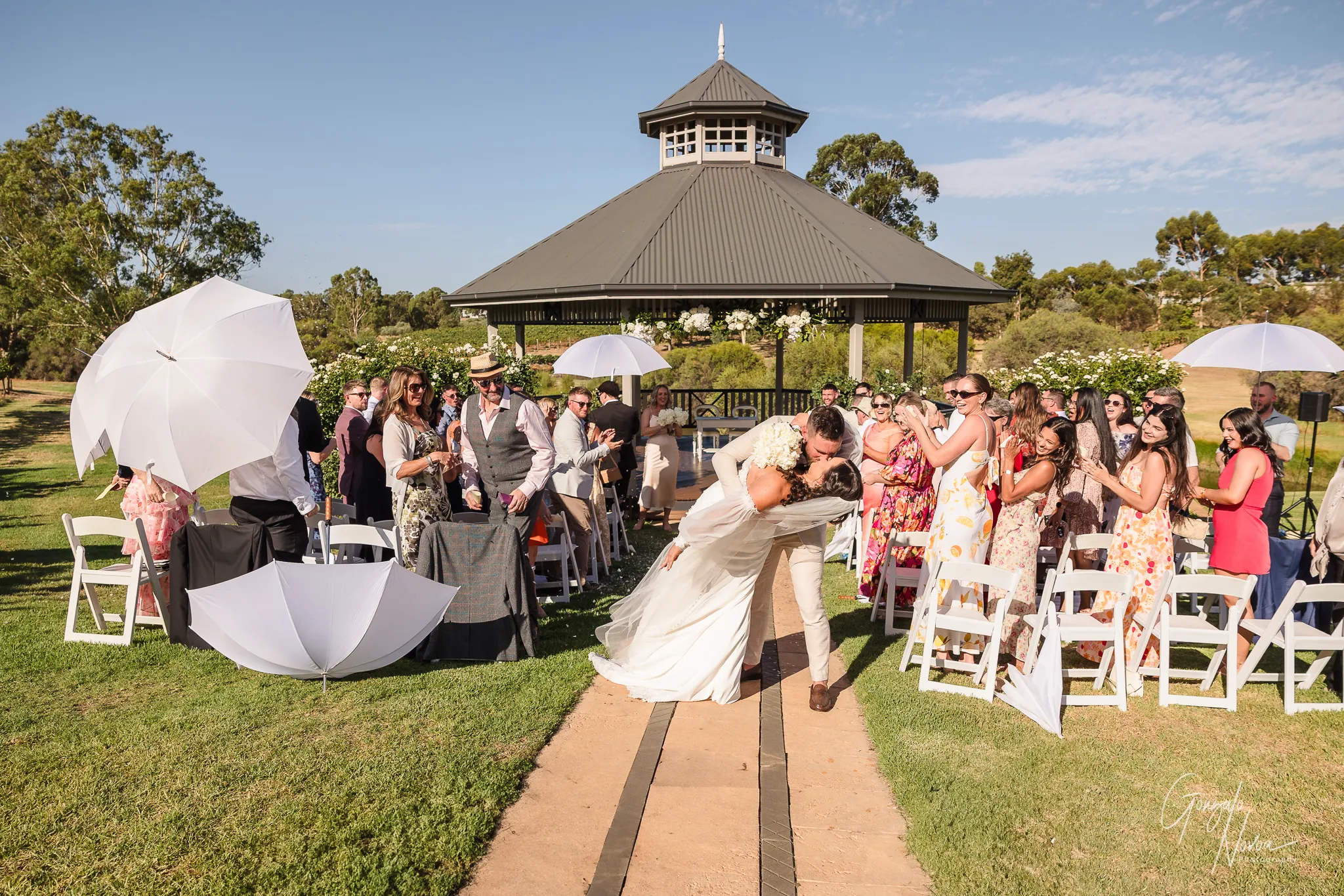 Bride and groom share a joyful dip kiss at the end of their wedding ceremony aisle, surrounded by cheering guests and white parasols under a sunny sky at Sittella Winery.
