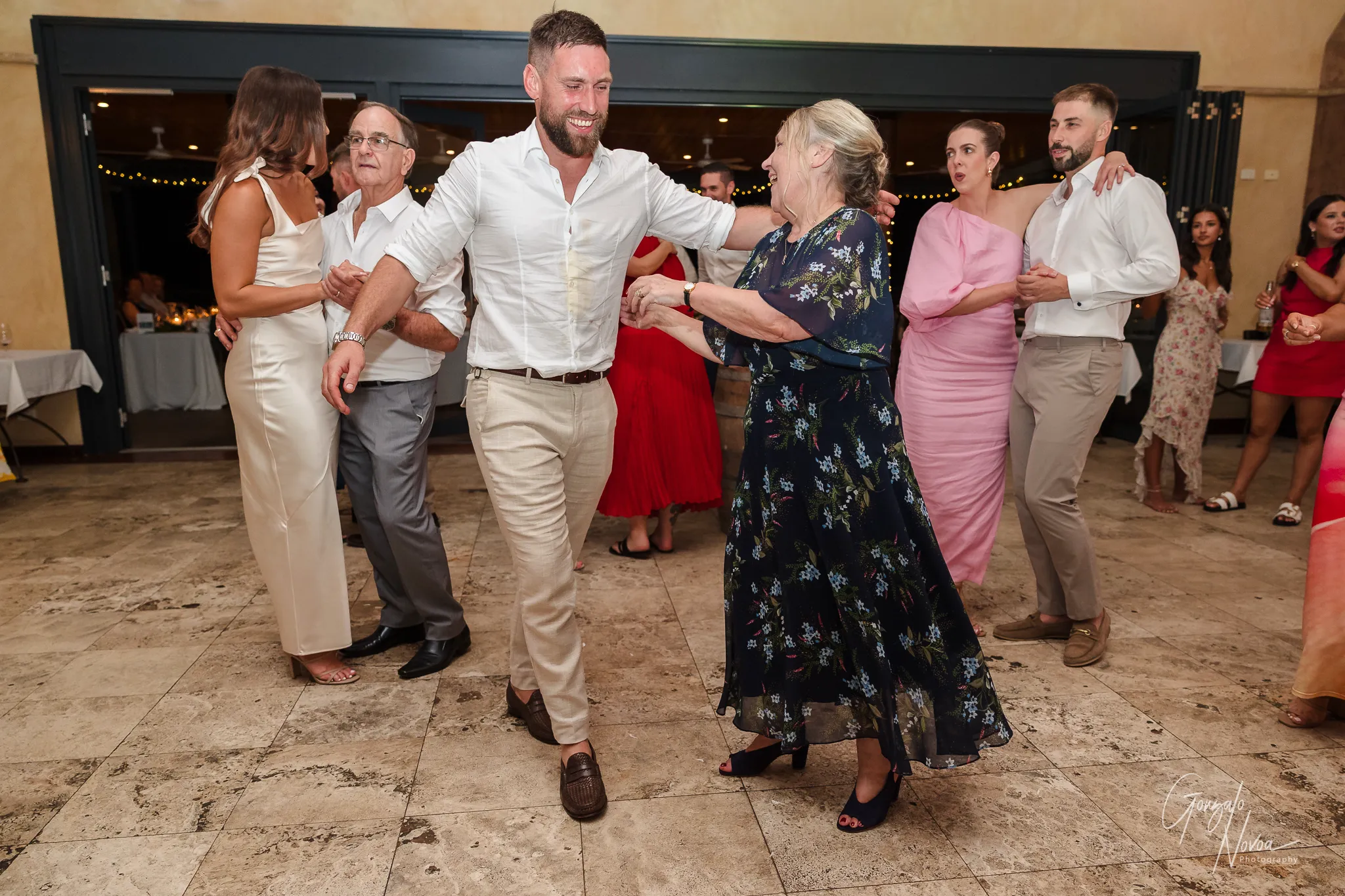 The bride and her father dance together during the wedding reception, laughing and holding hands in a lively, happy moment. The bride wears a strapless wedding gown with sheer off-shoulder sleeves, and her father is dressed in a white shirt and grey trousers. In the background, guests cheer them on, some taking photos and videos, while fairy lights add a festive touch to the evening atmosphere.