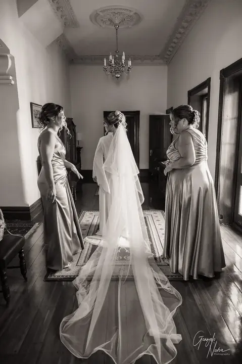 Bride with long veil standing in a hallway at Buckland Estate, surrounded by bridesmaids in satin dresses during wedding prep.