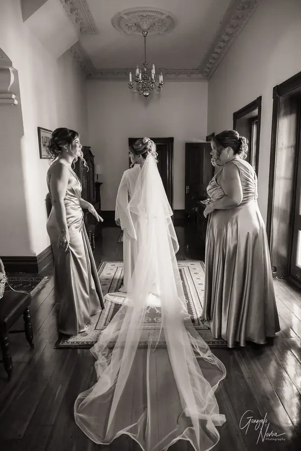 Bride with long veil standing in a hallway at Buckland Estate, surrounded by bridesmaids in satin dresses during wedding prep.