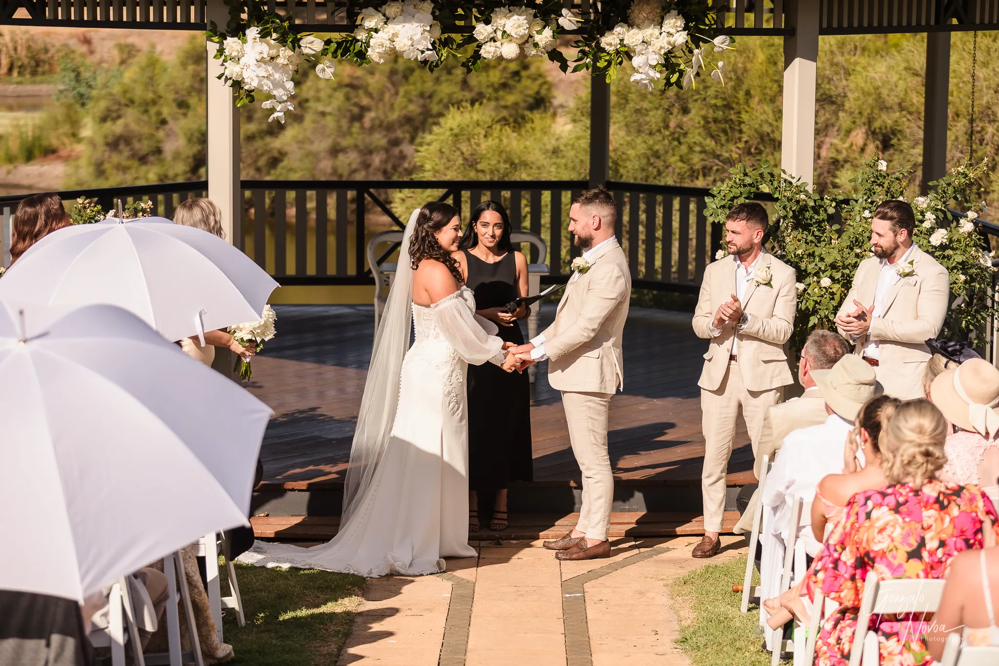 Bride Tahnee and groom James standing hand in hand at the altar of Sittella Winery’s gazebo, smiling at each other as their celebrant and wedding party look on under a floral canopy.