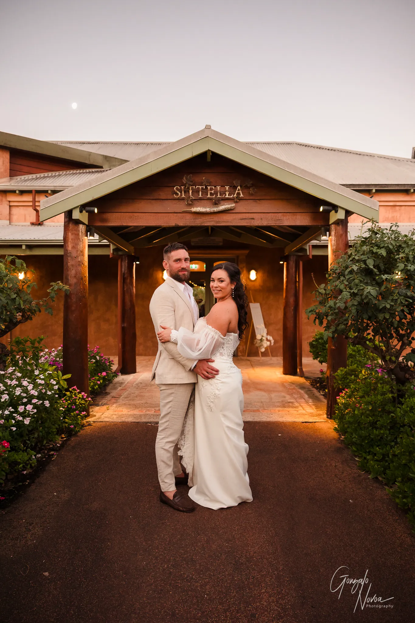 Bride and groom embracing under the entrance archway of Sittella Winery at sunset, with soft golden light illuminating the path and lush greenery on either side