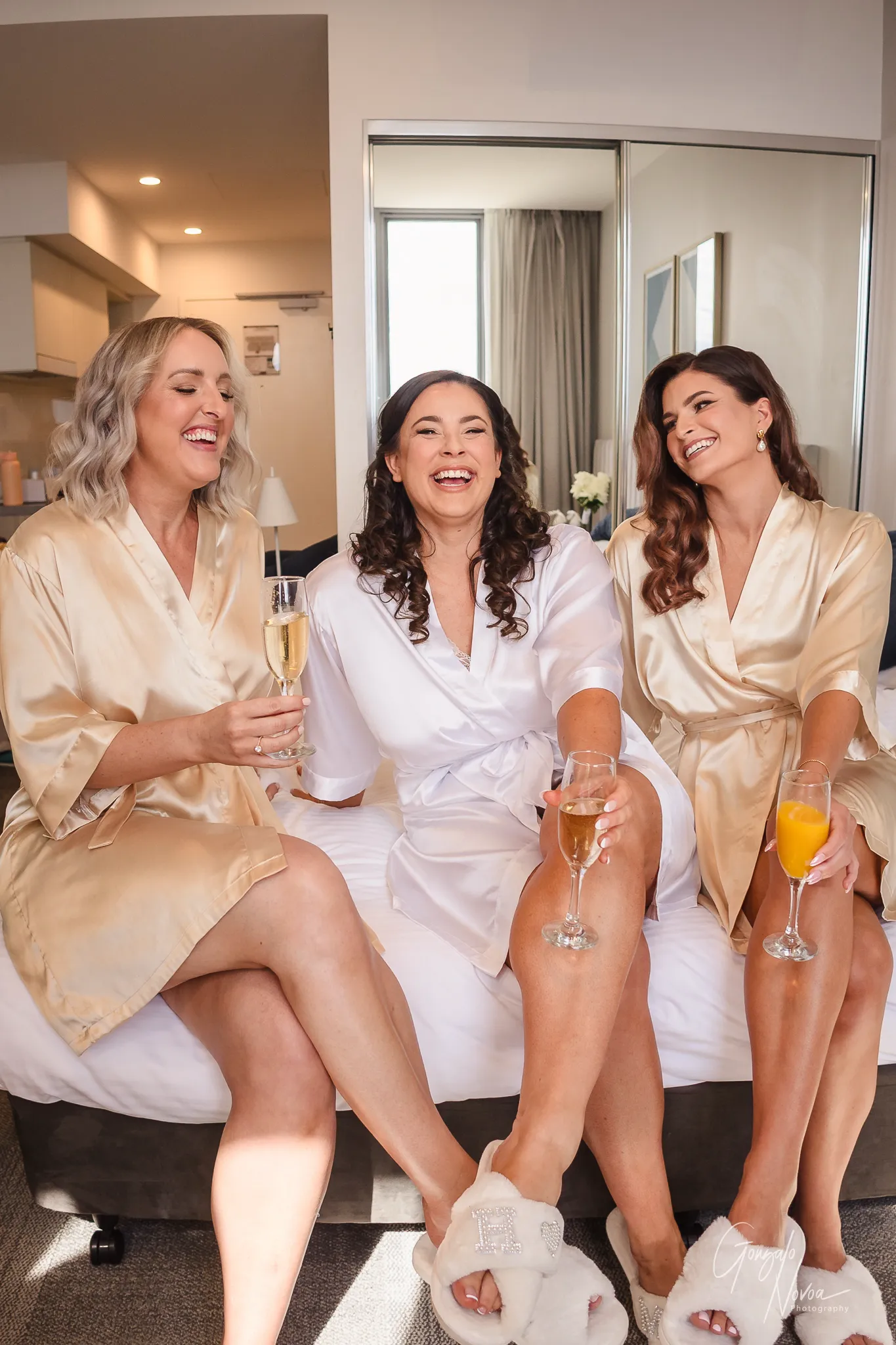 Tahnee and her two bridesmaids laughing together while sitting on a bed, wearing satin robes and holding champagne and orange juice glasses. The room is filled with morning light and joyful energy before the wedding ceremony.