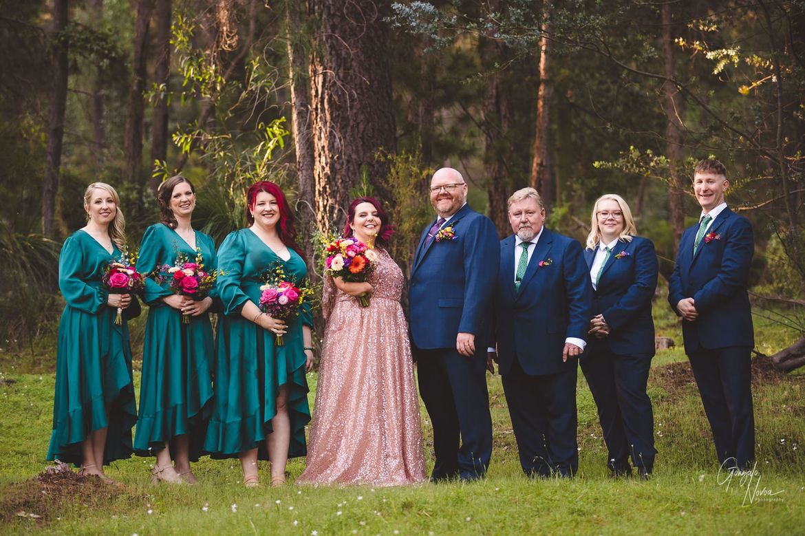 Sophie and Bradford with their wedding party near Mundaring Weir, Mundaring, Western Australia. The bride is in a sparkling pink dress, the groom in a blue suit, and the bridesmaids in teal dresses holding vibrant bouquets. The groomsmen are in matching blue suits, creating a colourful and joyous scene against the natural backdrop.