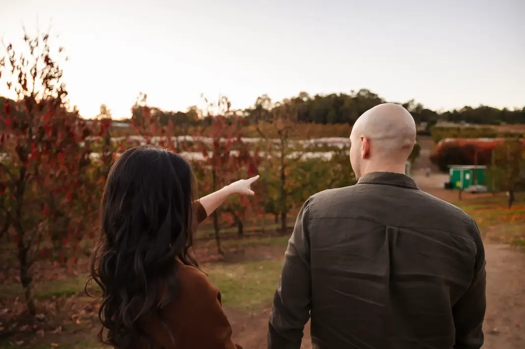 Sandra pointing across the orchard while standing beside Luis, both facing away from the camera. They’re surrounded by autumn trees with shades of red and green, overlooking a quiet farm path.