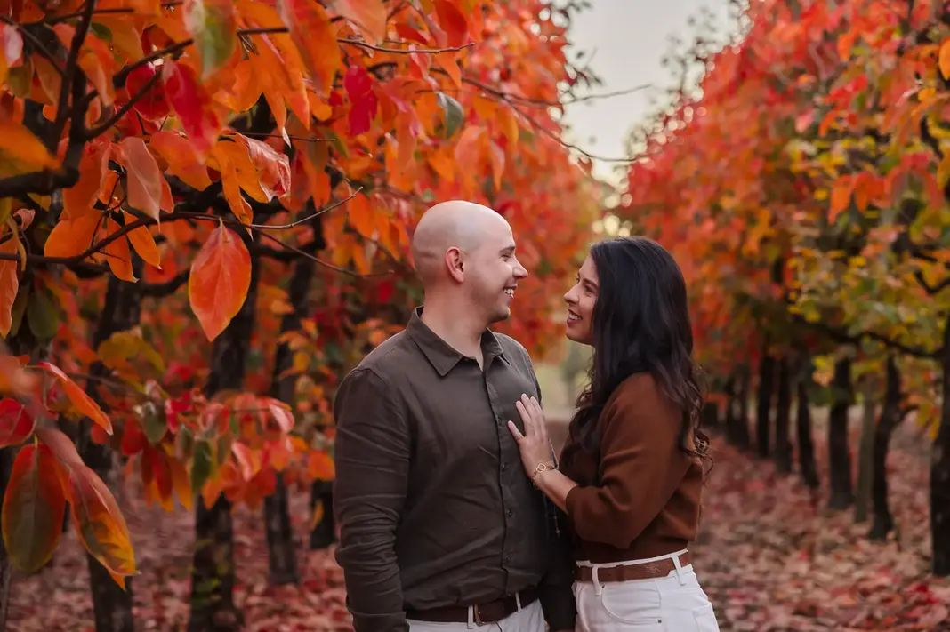 Sandra and Luis standing close together, smiling at each other beneath vibrant autumn foliage at Raeburn Orchards. Red and orange leaves surround them, with soft light filtering through the trees.
