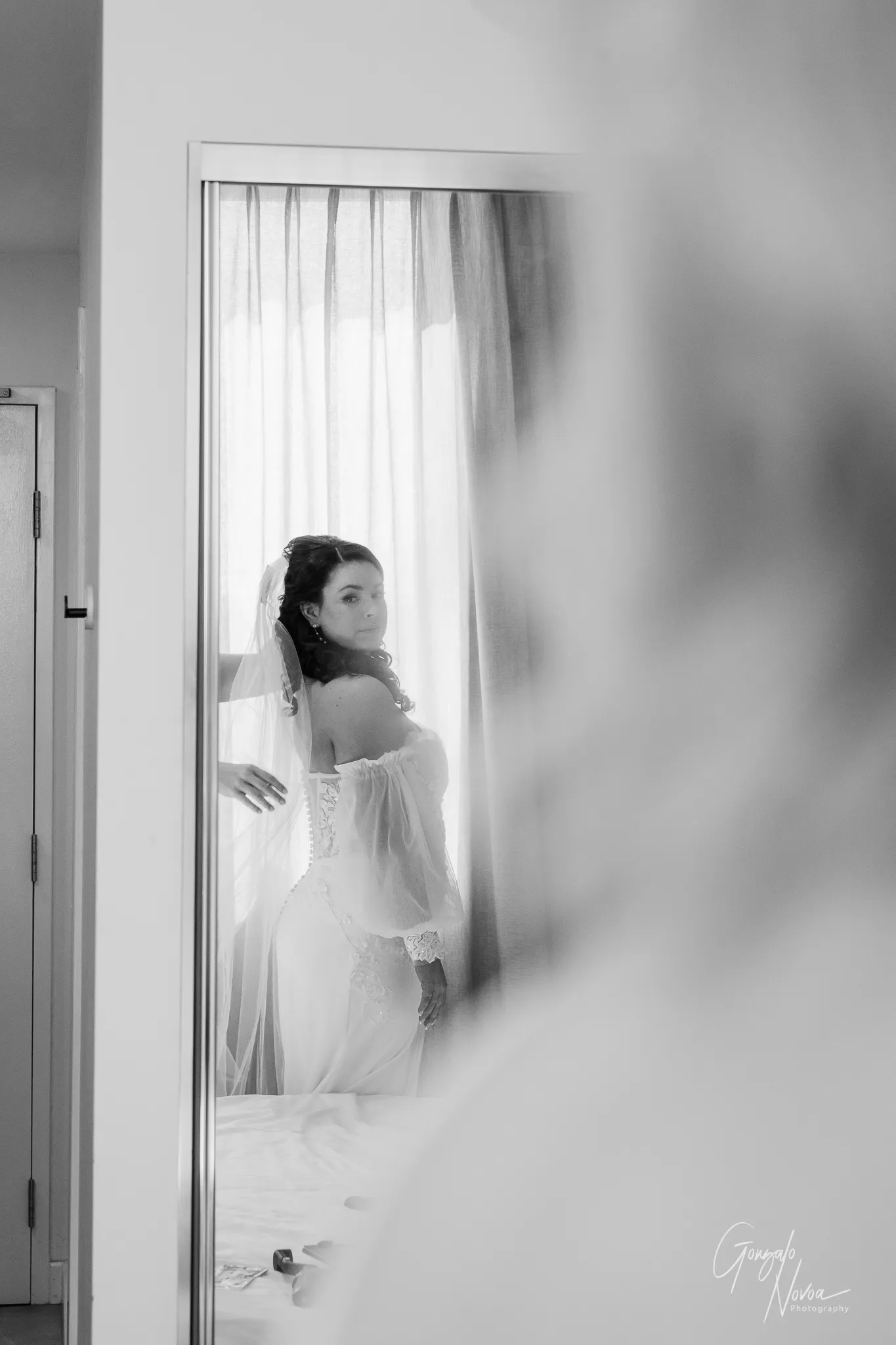 Black and white photo showing Tahnee’s reflection in a mirror as she stands near a bright window. She is dressed in her lace wedding gown, adjusting her veil and soaking in the emotion before the ceremony.