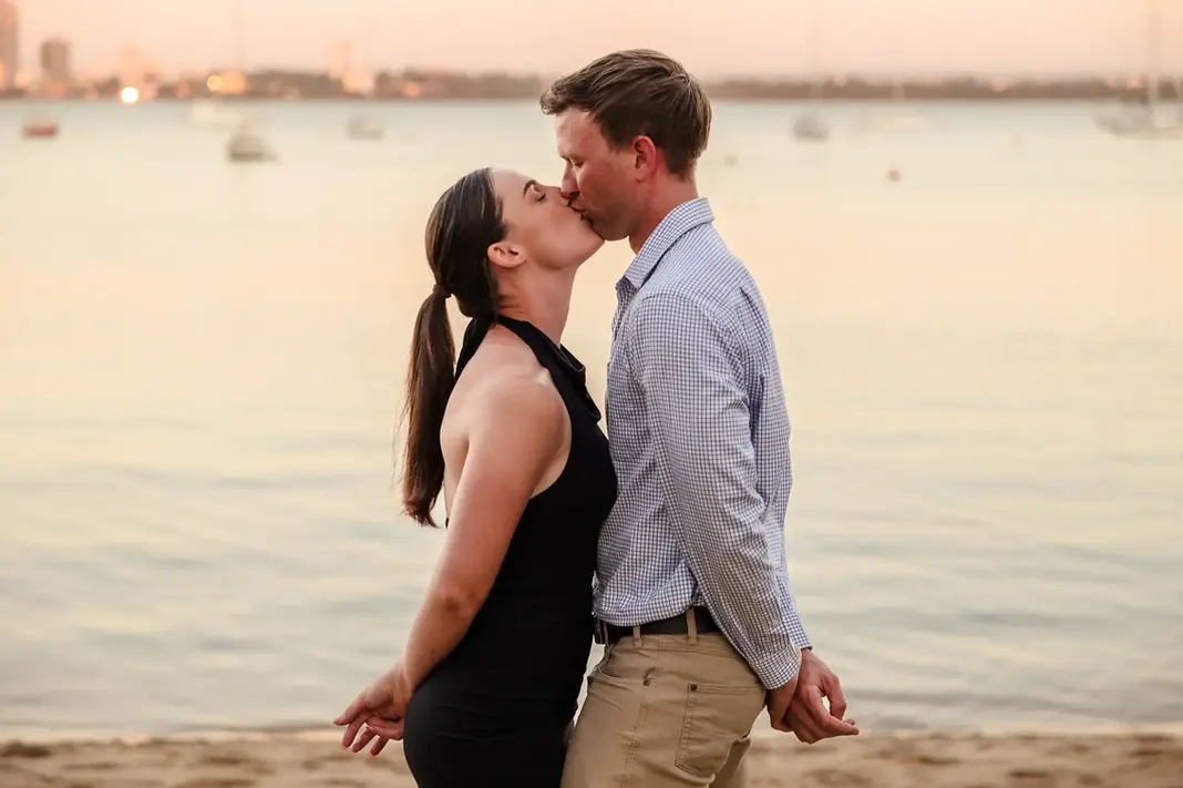 Baleigh and Kailym kissing on the beach at Matilda Bay Reserve at sunset, holding hands behind their backs with soft water and boats in the background.
