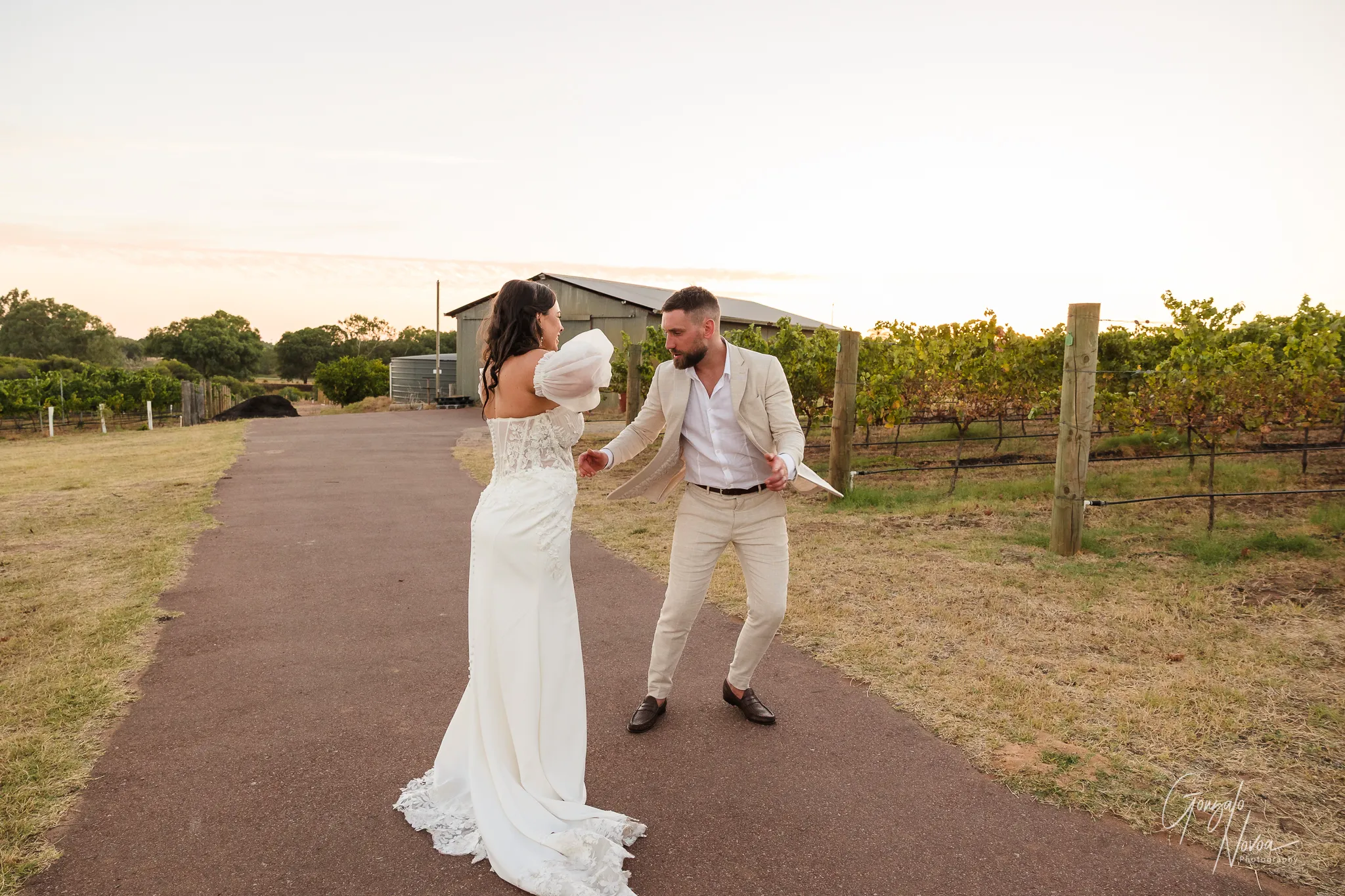 Bride and groom laughing and dancing together on a paved path surrounded by vineyards at sunset, with the groom playfully crouched in front of the bride who is smiling and lifting her arms.