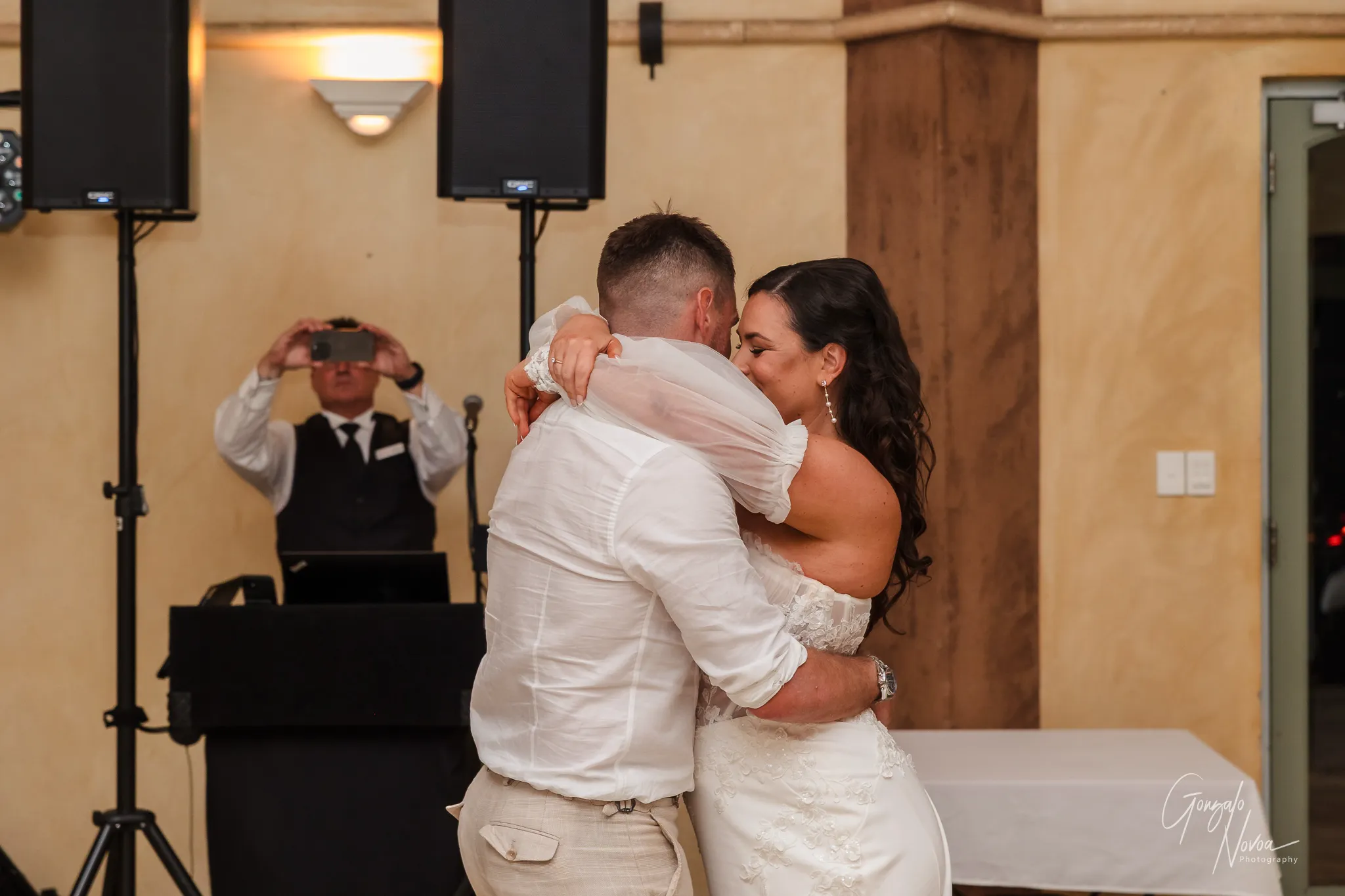 The bride and groom share an emotional embrace during their first dance at the reception. The bride smiles with her eyes closed, resting her head on the groom’s shoulder, while he holds her tightly. She wears an off-shoulder lace gown with delicate sheer sleeves, and he is dressed in a white shirt and beige trousers. In the background, a staff member is capturing the moment on a mobile phone.