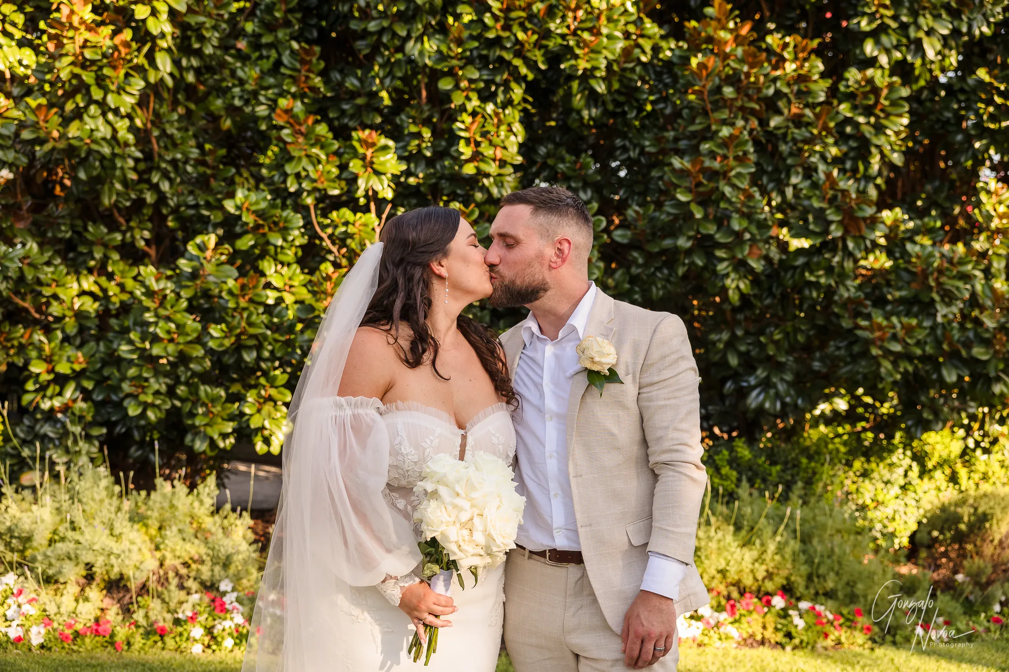 Tahnee and James share a kiss in front of lush greenery, Tahnee holding her bouquet and James gently leaning in
