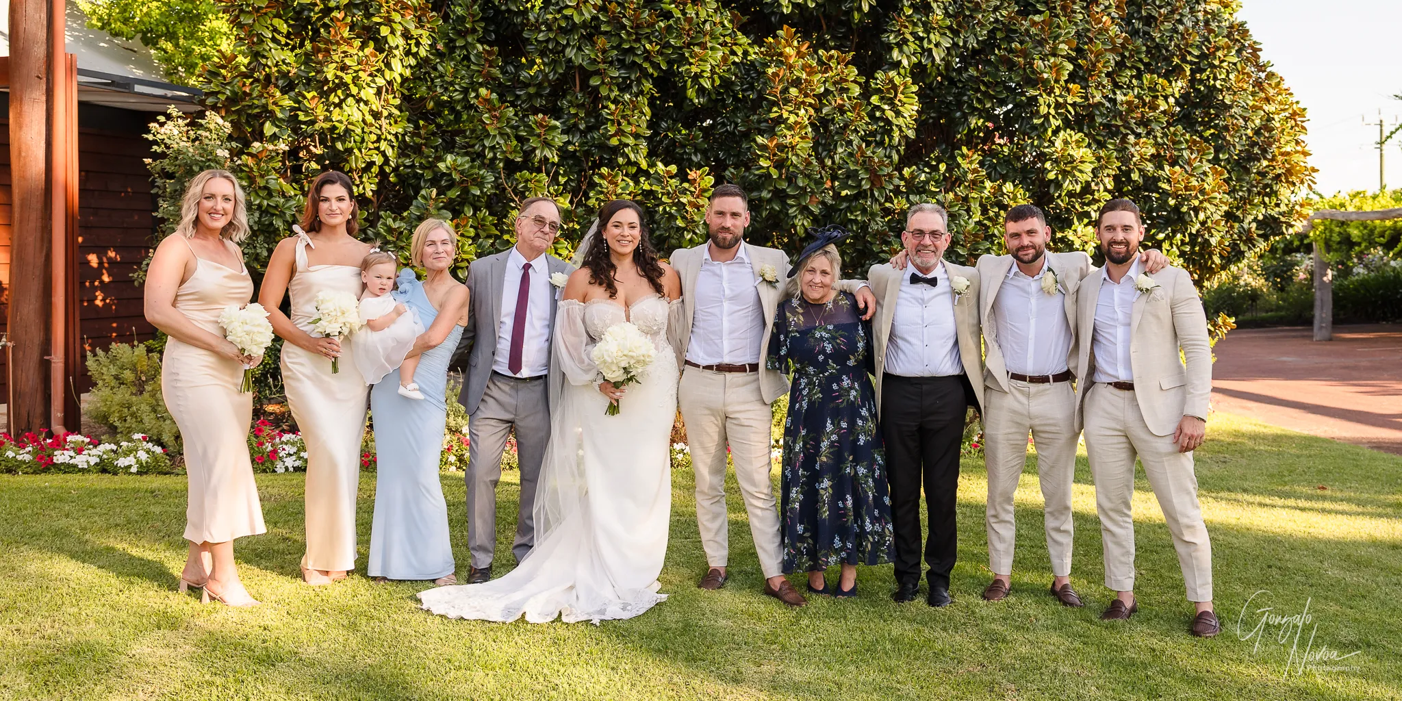 Tahnee and James standing with their bridal party and both sets of parents, smiling for a group portrait in front of a large tree and garden backdrop