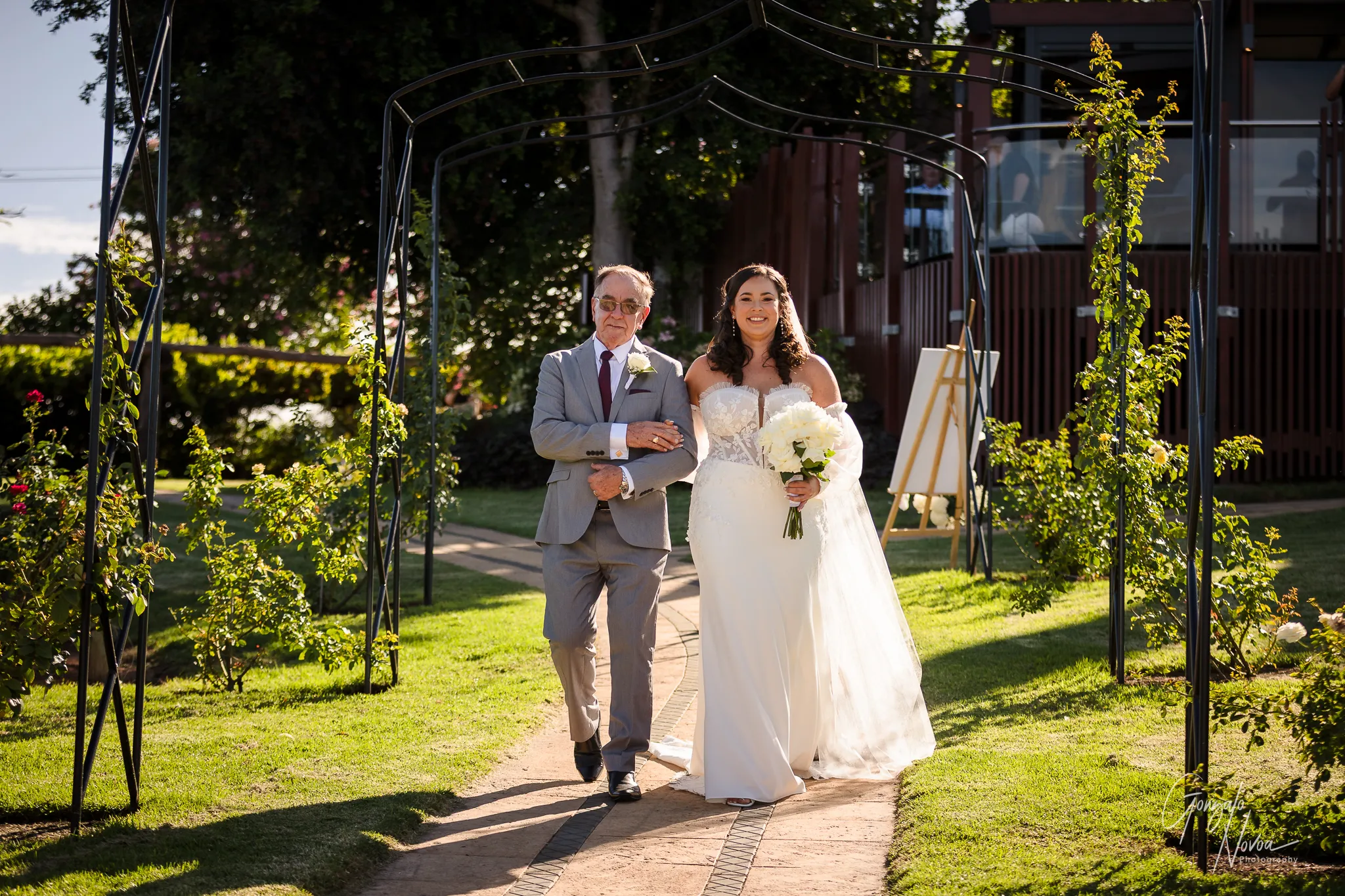 Tahnee smiles radiantly as she walks arm-in-arm with her father down the vine-lined path toward the ceremony at Sittella Winery, bathed in golden afternoon light.