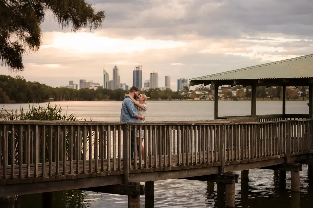Faith and Alex embracing on a wooden boardwalk over Lake Monger with the Perth city skyline and cloudy sunset sky in the background.