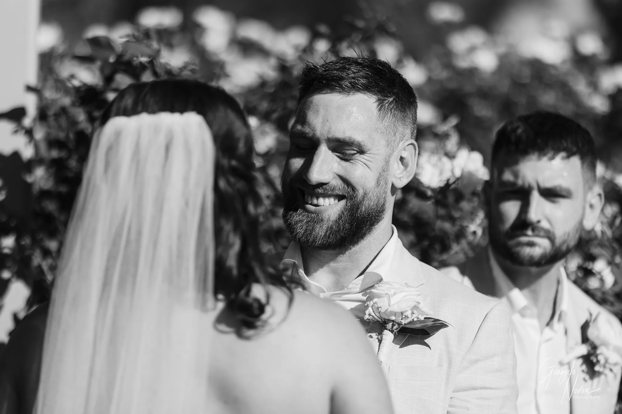 Black and white photo of groom James smiling warmly at bride Tahnee during their wedding ceremony, with his best man watching emotionally in the background.